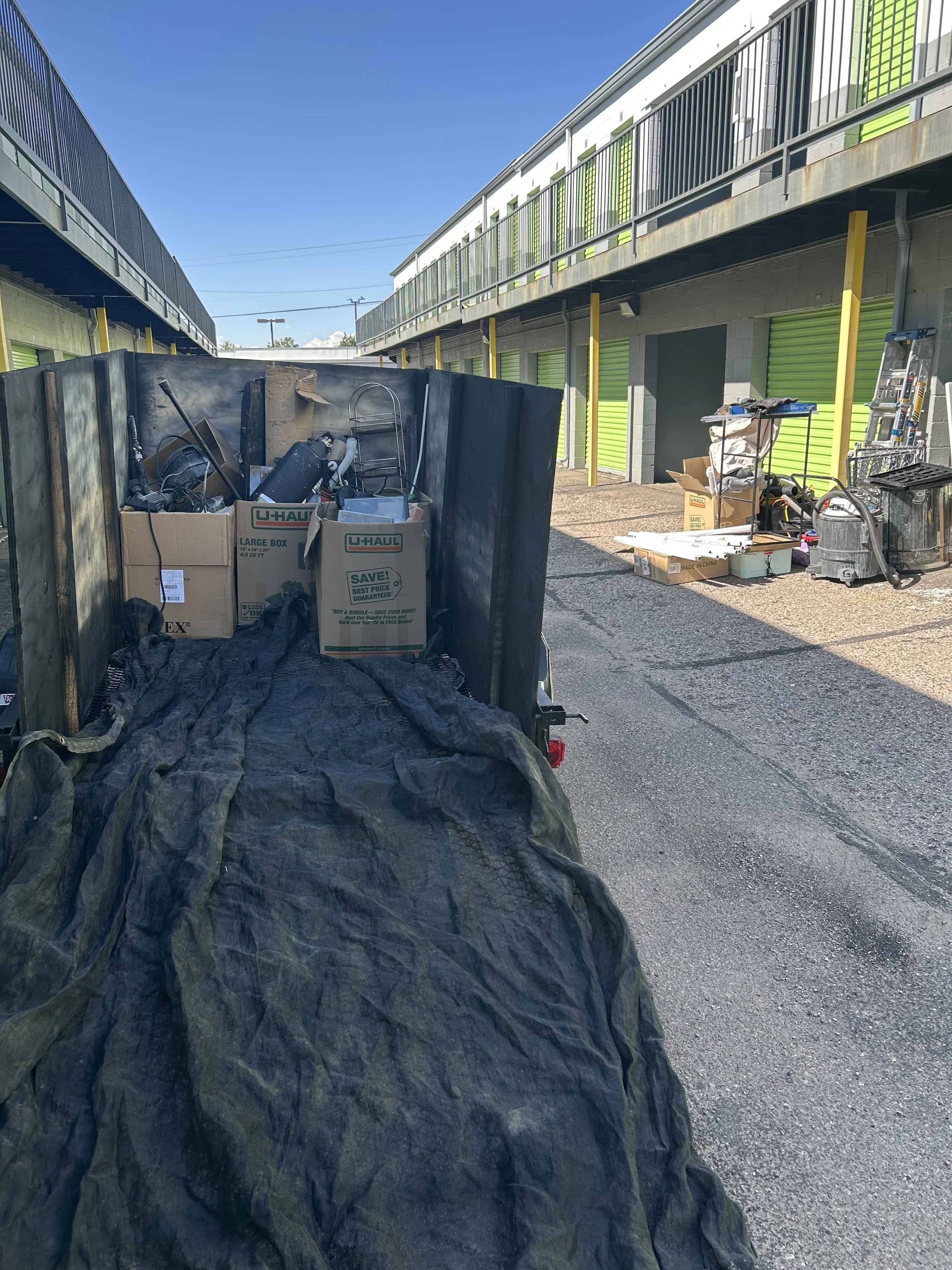 View of an outdoor storage or parking area with multiple storage units with green doors, a trash bin with miscellaneous items, and a black tarp covering some objects in the foreground. Clear blue sky overhead.