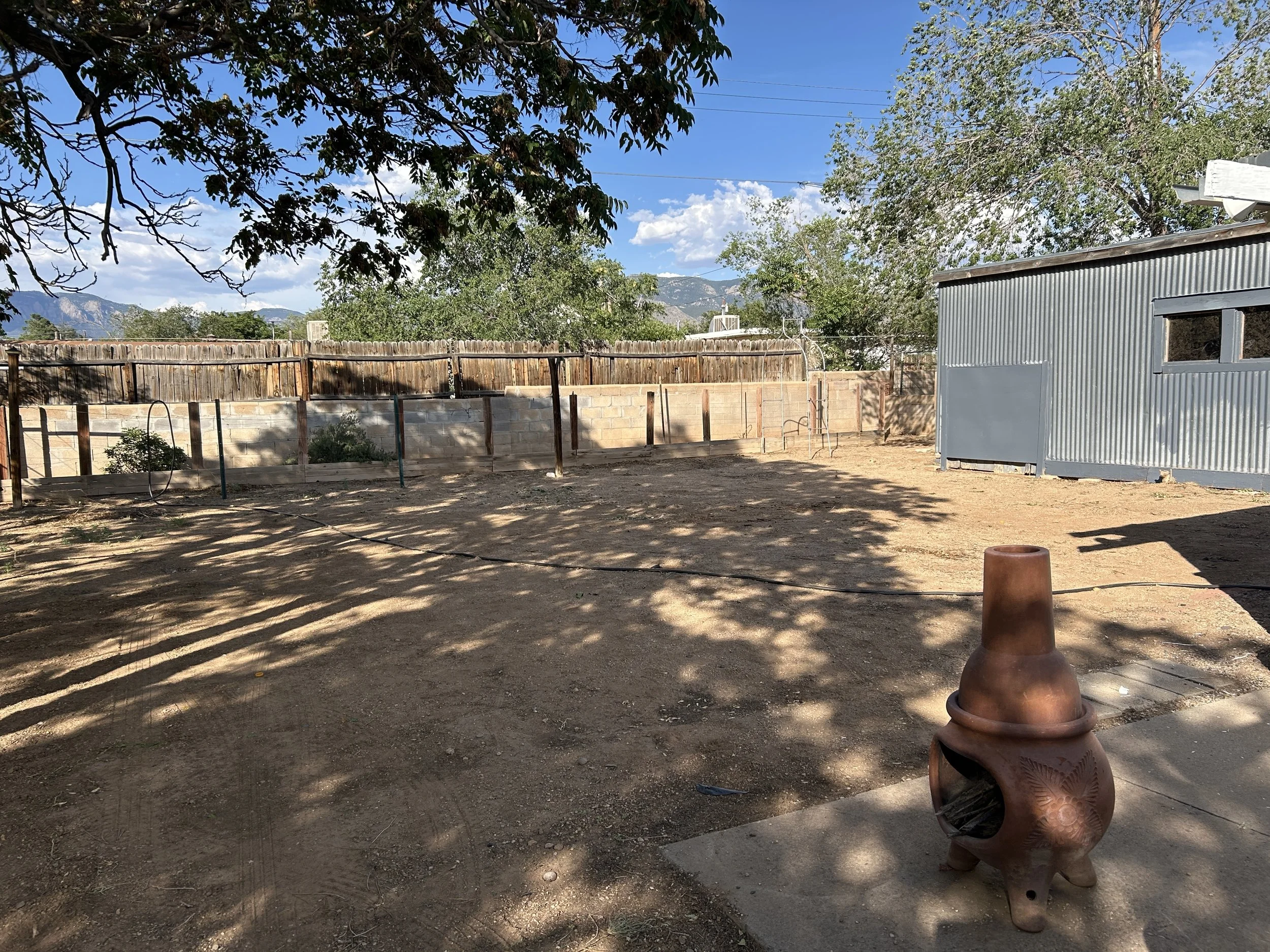 A backyard scene with a metal shed, a wooden fence, and trees. Mountains are visible in the background under a blue sky with some clouds. Shadows from the trees are cast on the ground, and there is a large clay pot in the foreground.