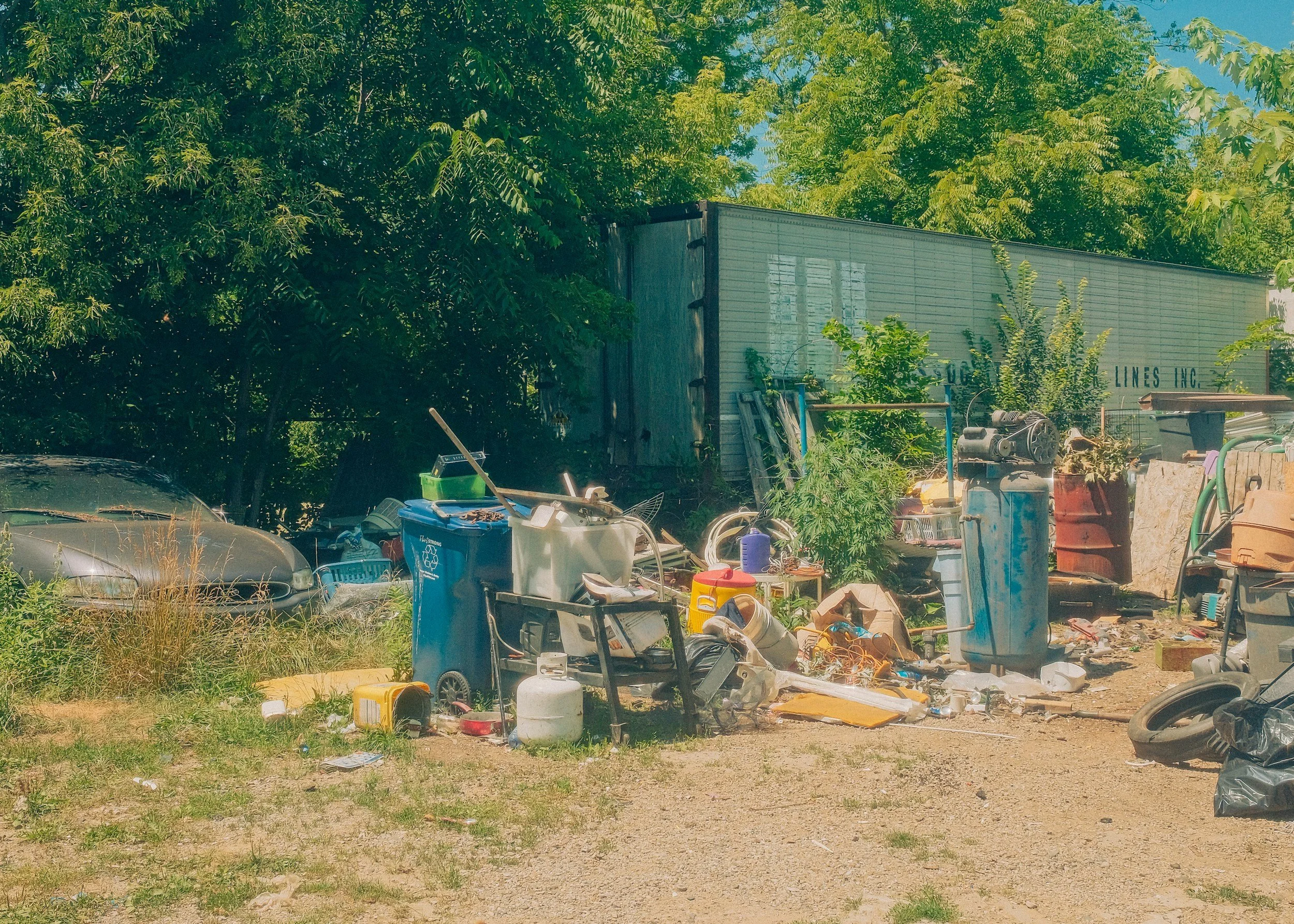 A cluttered outdoor area with trash, discarded items, and various objects in front of trees and a building.