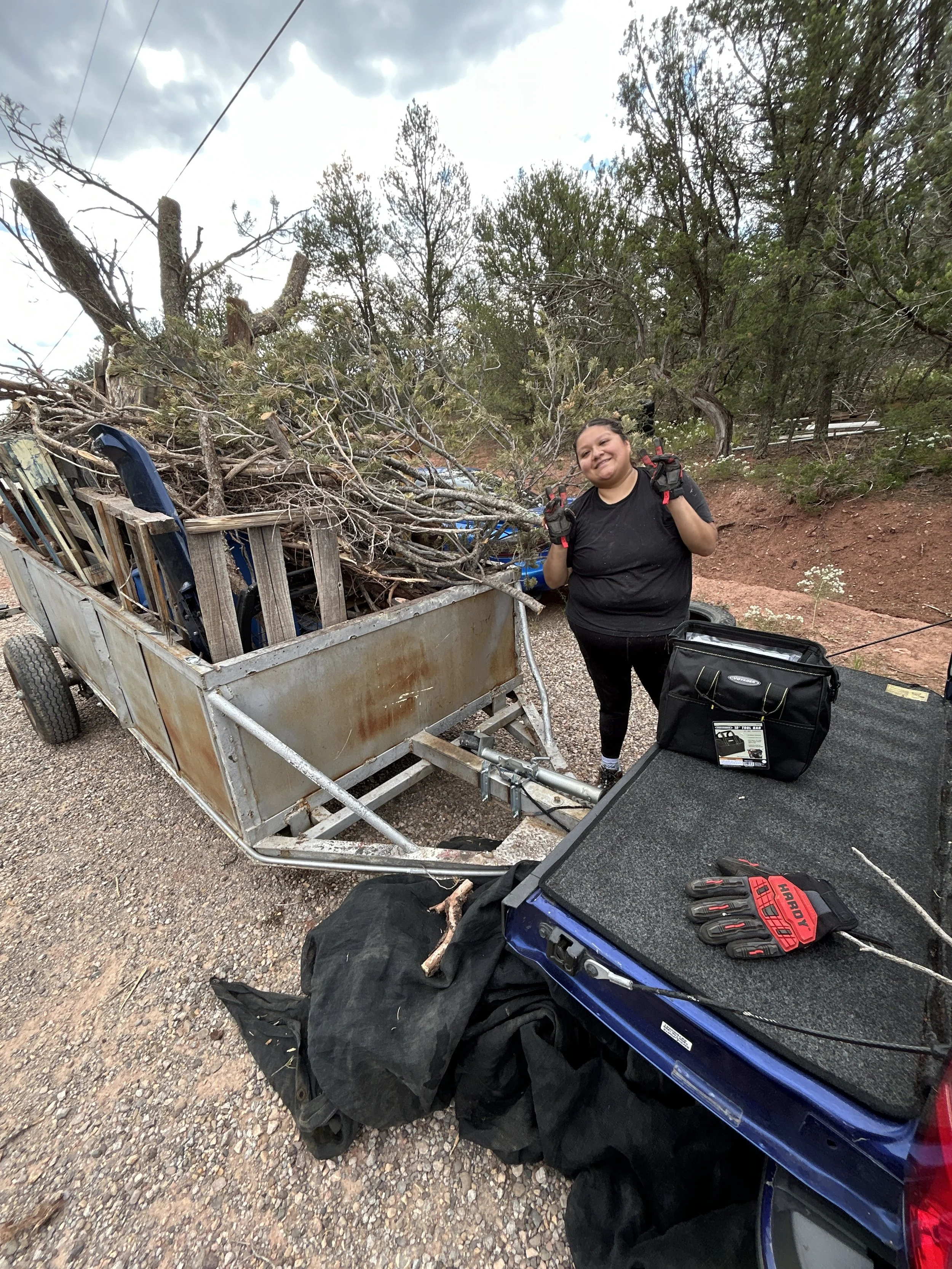 A woman standing next to a trailer filled with fallen tree branches. She is wearing gloves, a black t-shirt, and has a cheerful expression. There are trees and cloudy sky in the background, and tools are on a table nearby.