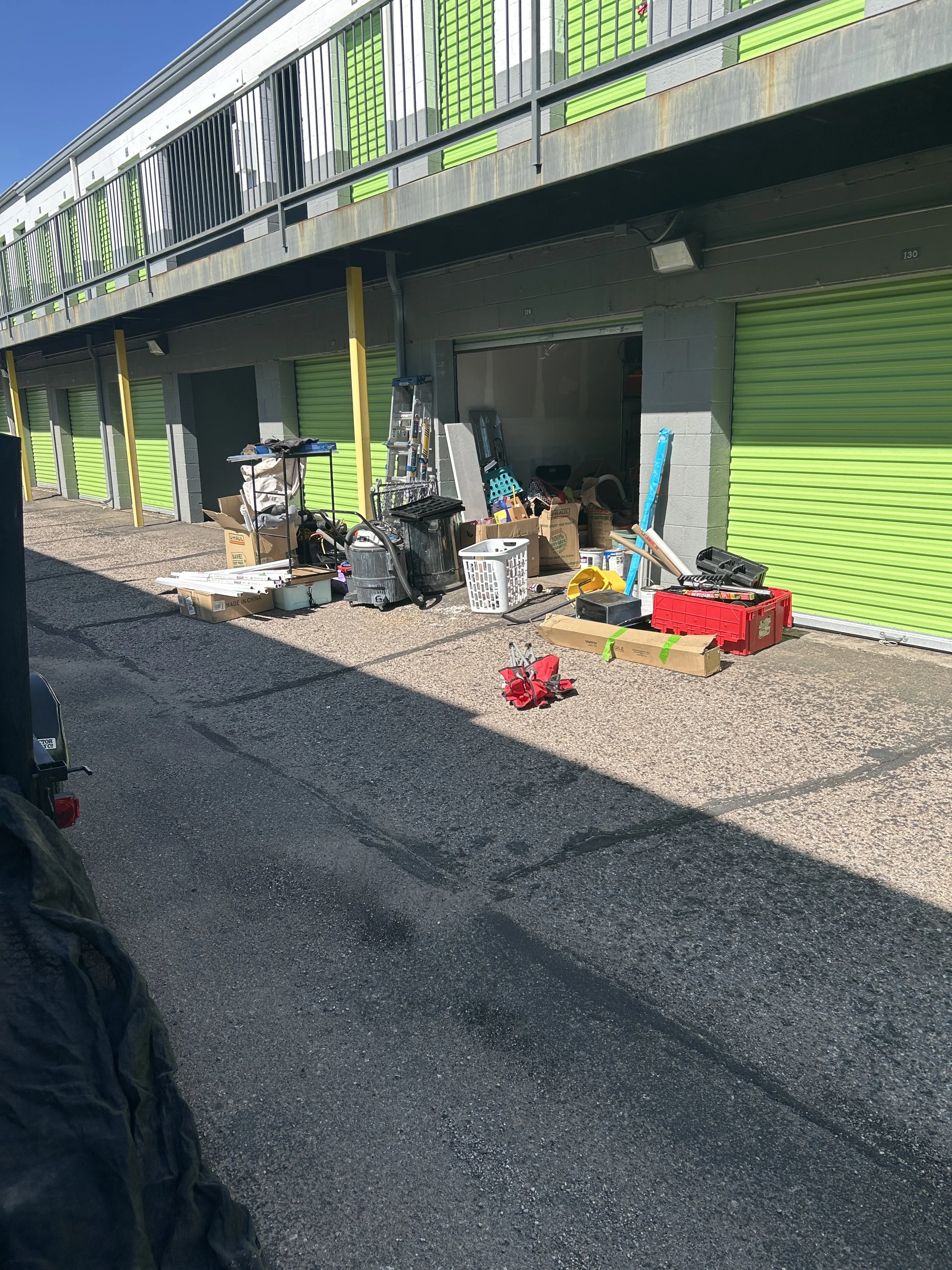 An outdoor storage unit area with bright green roll-up doors, items like boxes, tools, and household supplies placed outside, and a concrete parking lot in front.