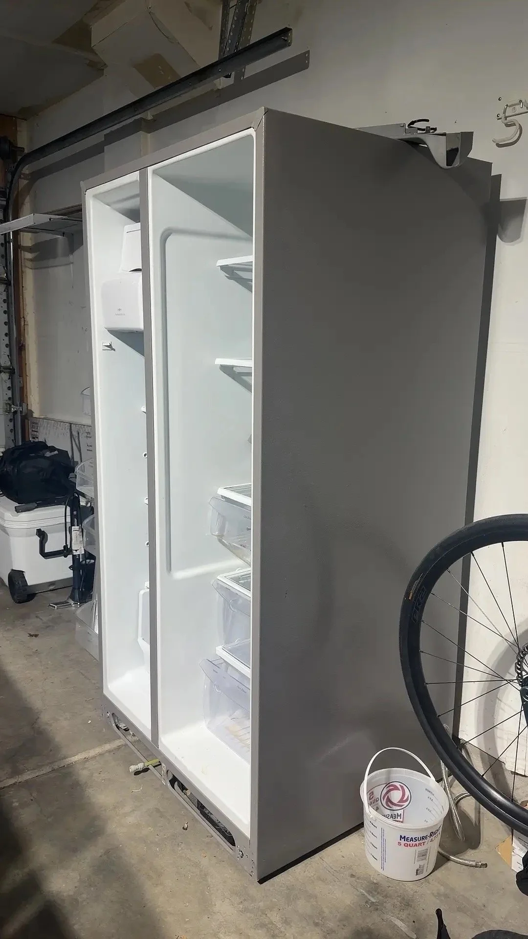 Empty white refrigerator in a garage, partially open with transparent storage bins on the shelves.