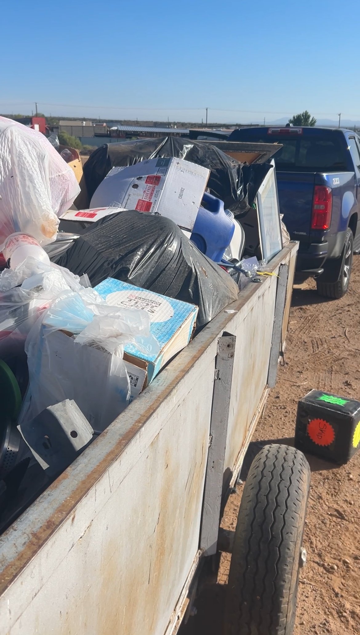 A utility trailer filled with household trash and discarded items, attached to a vehicle in an outdoor setting.