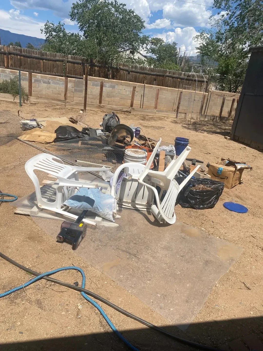 Cluttered backyard with overturned white plastic chairs, various debris, black trash bags, a blue bucket, a cardboard box, and tools, with a wooden fence and trees in the background under a partly cloudy sky.