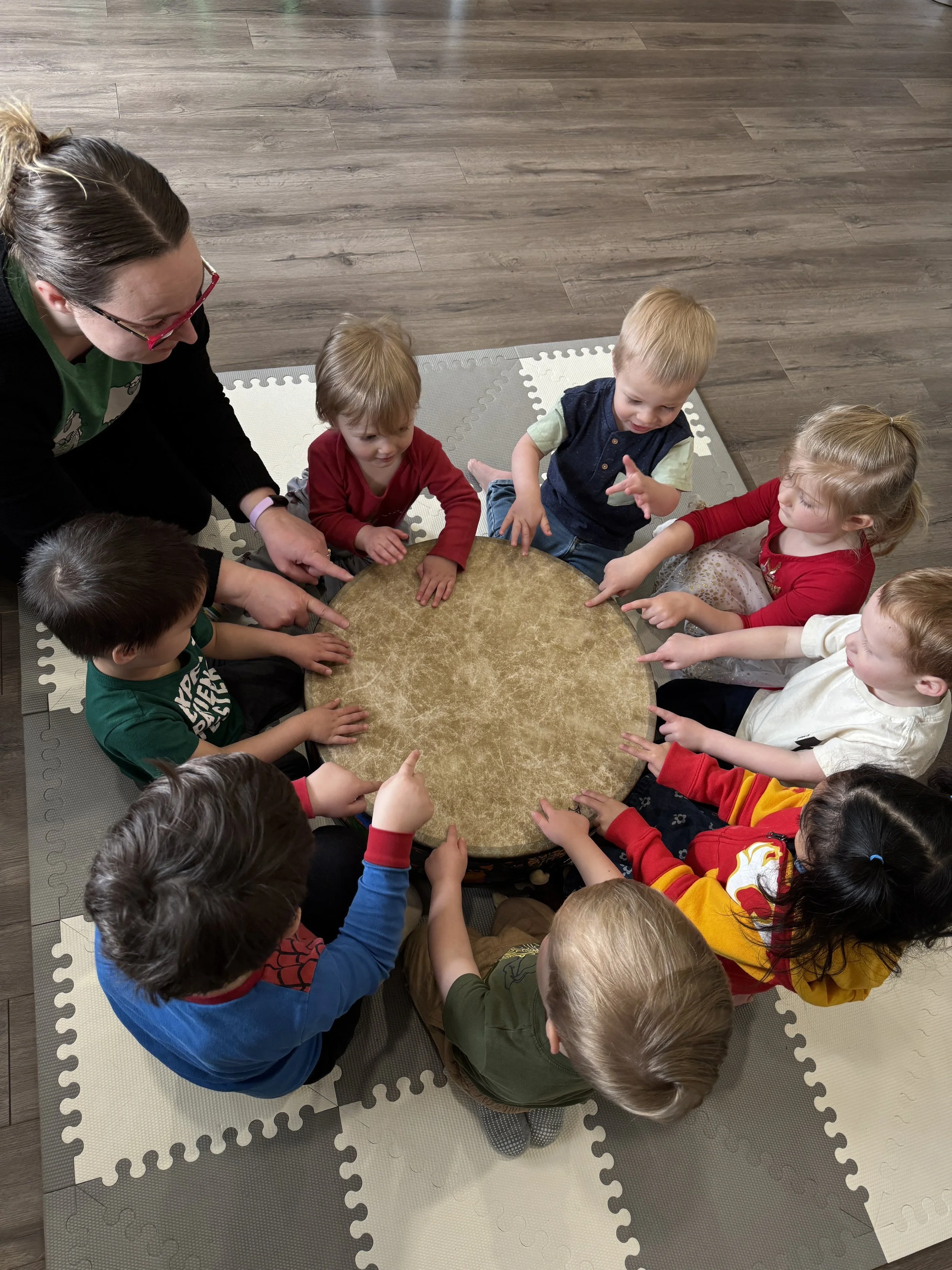 A group of young children sitting around a round table on a foam mat, engaged in a discussion led by Meagan Pon