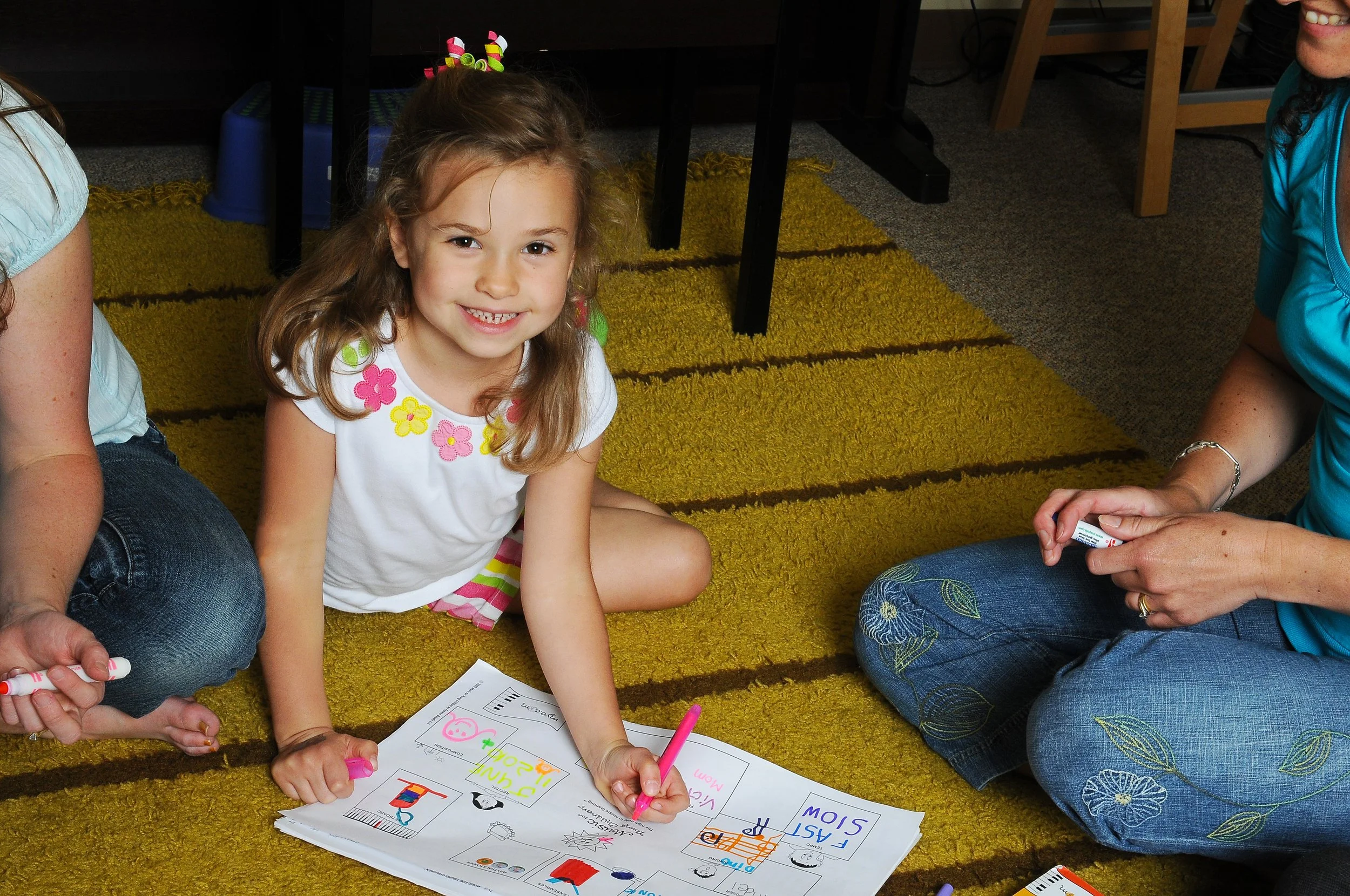 A young girl with light brown hair and a colorful hair accessory sits on a yellow rug, smiling at the camera while coloring on a worksheet with pink and orange markers. There are adults and other children nearby, part of a group activity in a classroom setting.
