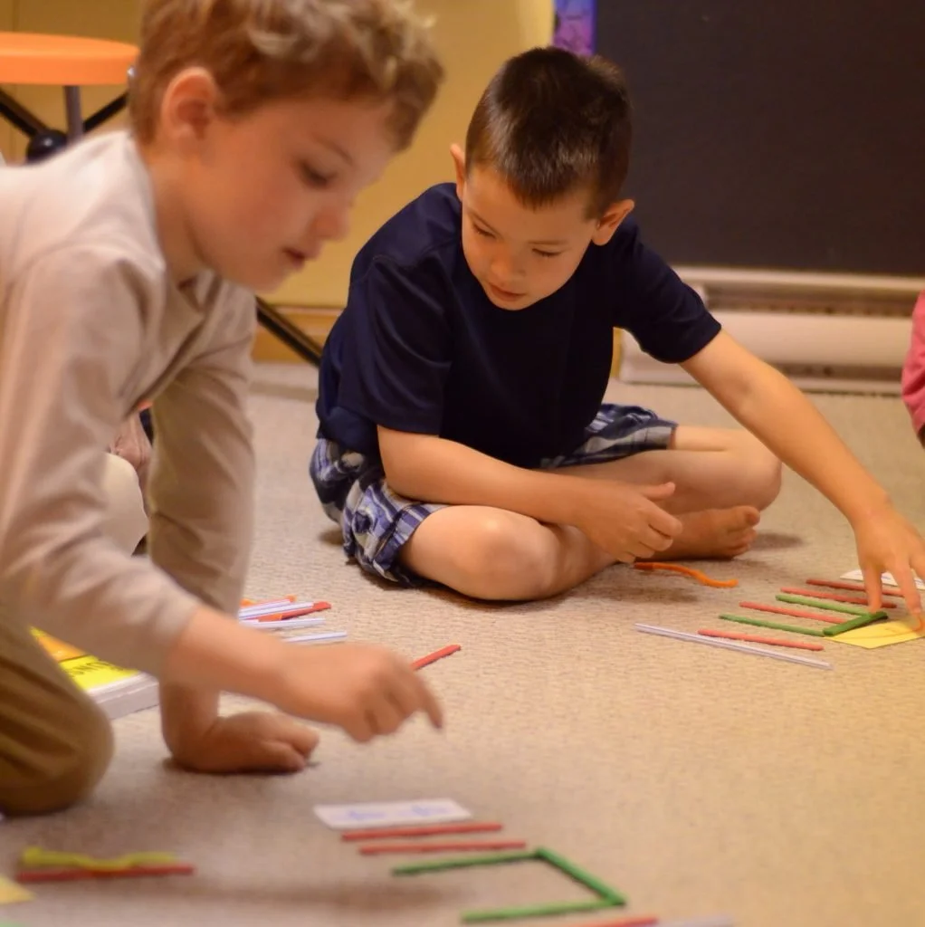 Two young boys sitting on the floor playing a game with colorful pipe cleaners and small cards.
