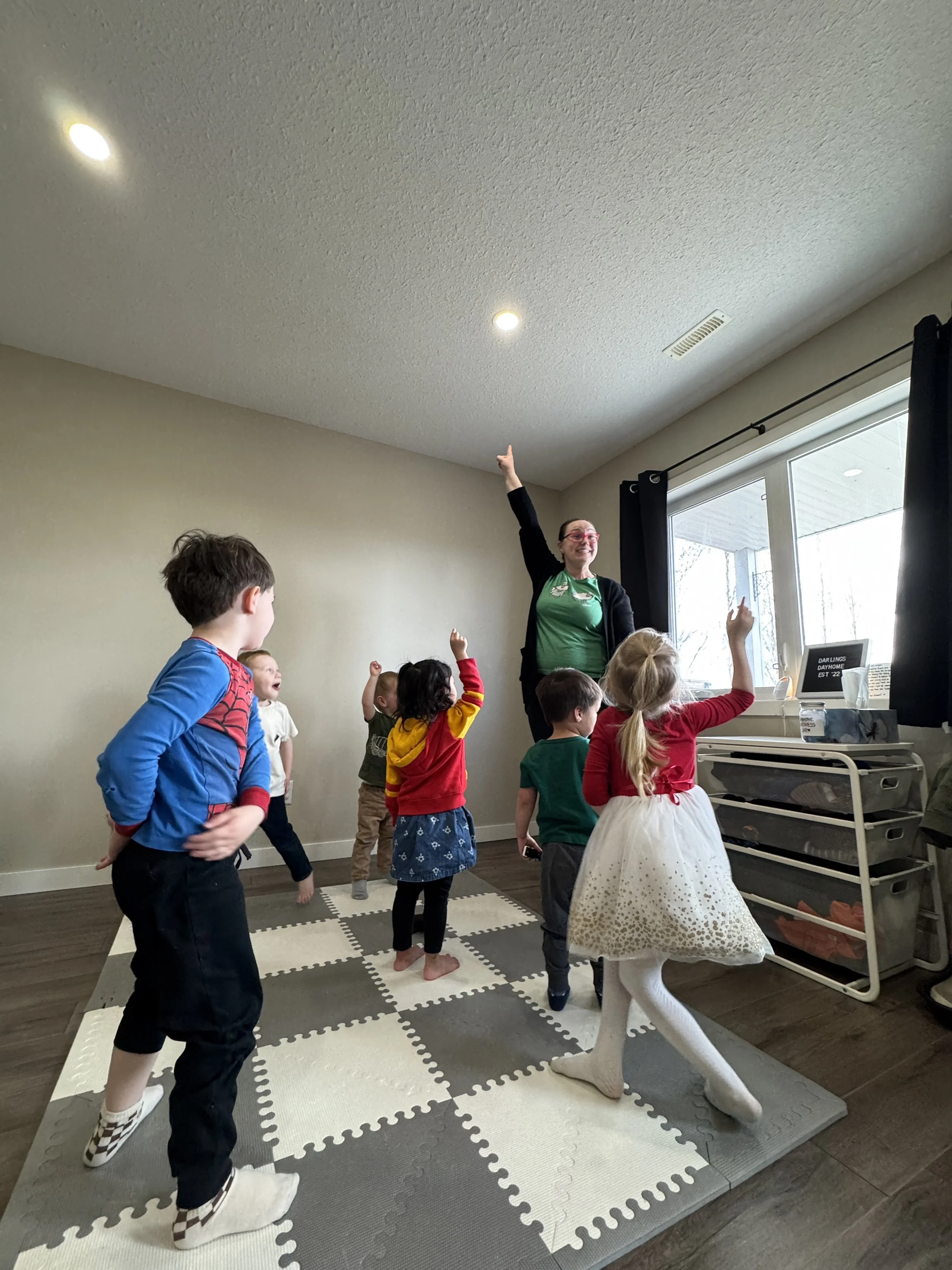 A group of children in a room with Meagan leading an activity. The children are raising their hands, and Meagan is pointing upward. They are playing musical games in Cochrane alberta.
