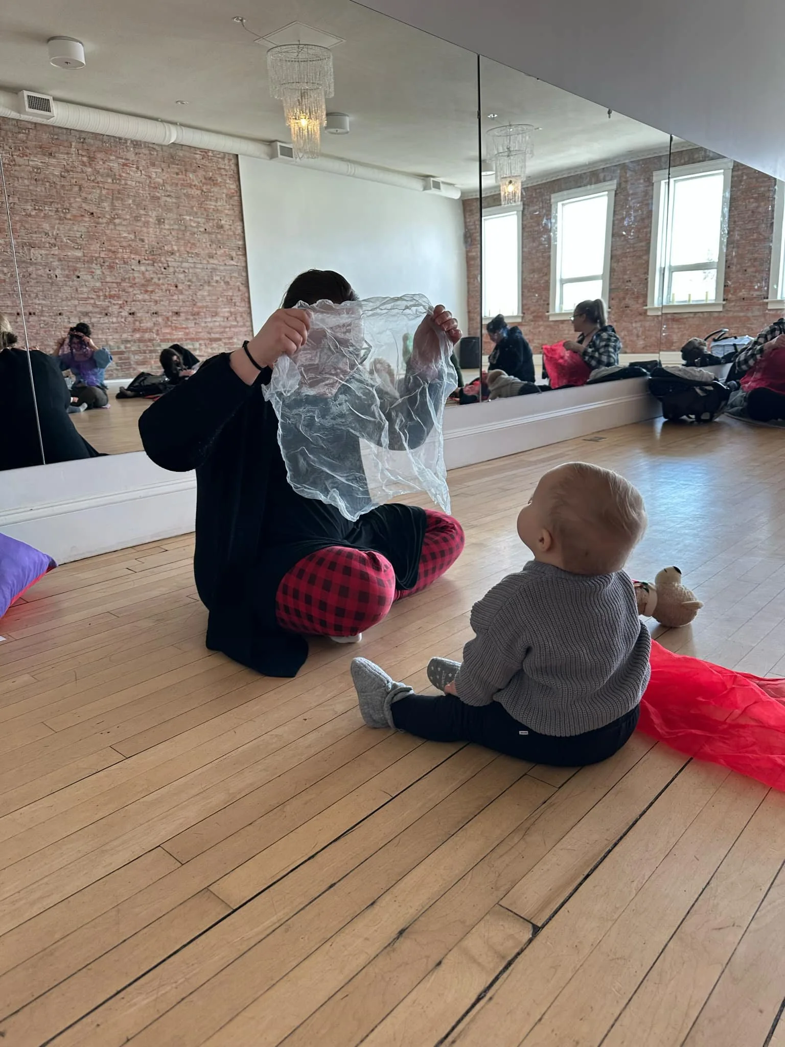 A group of children and a teenager sitting on a wooden floor in a dance or yoga studio with large windows and an exposed brick wall. One teenager is holding a piece of sheer fabric, and a child sitting in front of them looks at the fabric. Bags and belongings are scattered around the room.