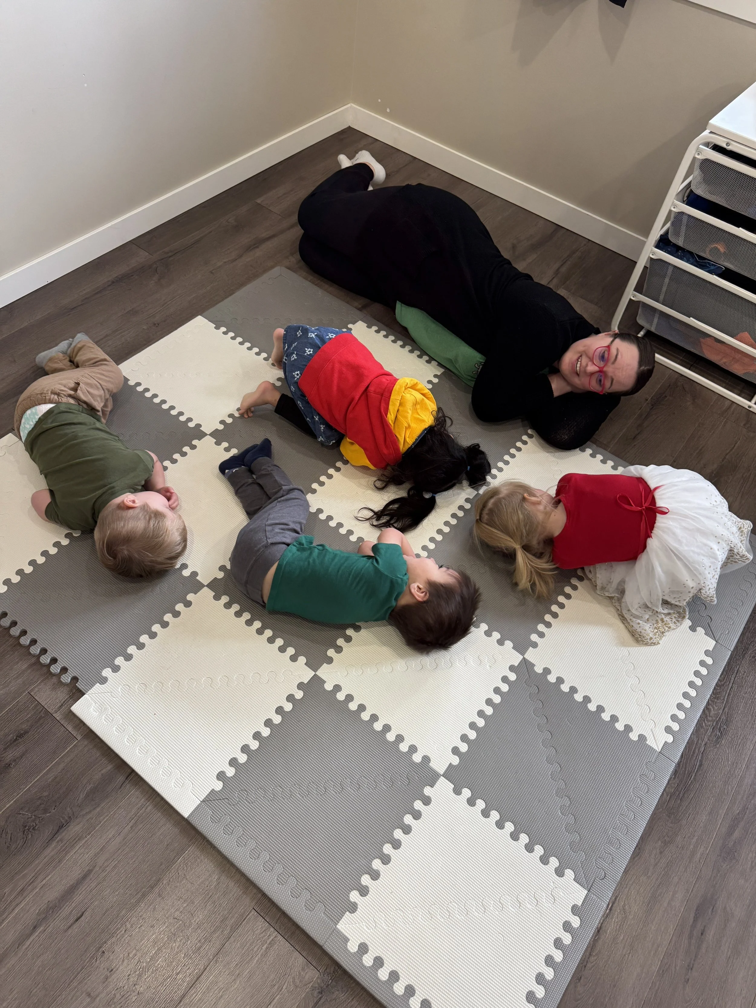 Meagan Pon dressed in black lying on a green pillow on a foam puzzle mat, surrounded by children, in a room with gray walls and wooden floors. . They are playing musical games in Cochrane alberta.