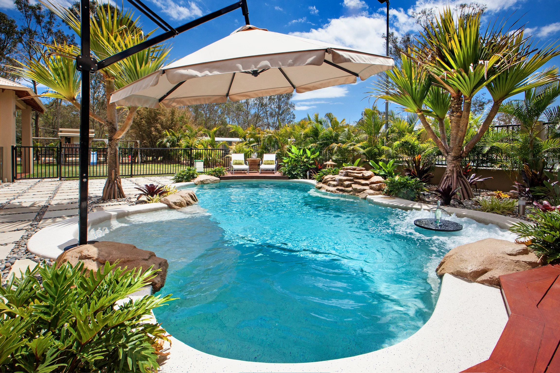 A backyard swimming pool surrounded by plants and trees under a blue sky with scattered clouds, with a large white patio umbrella overhead.