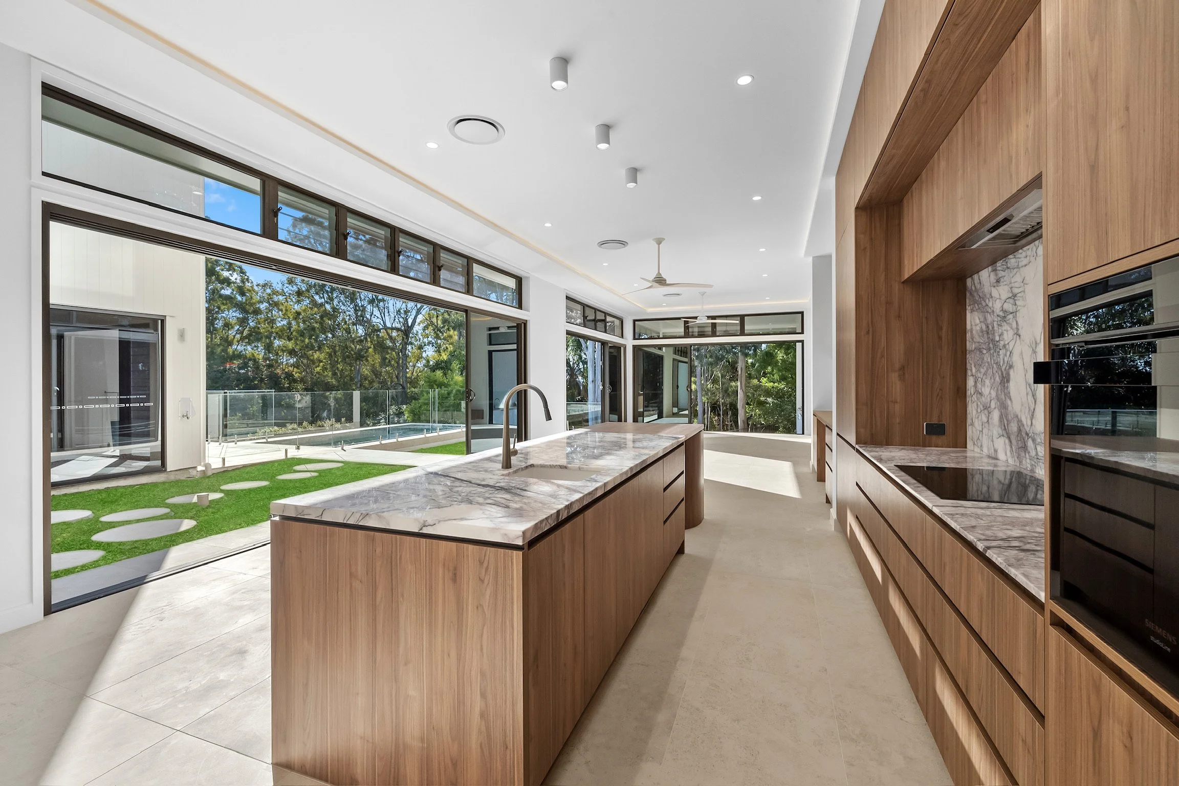 Modern kitchen with marble countertops, wooden cabinetry, large glass sliding doors revealing a backyard with patio and trees, and ceiling lights.