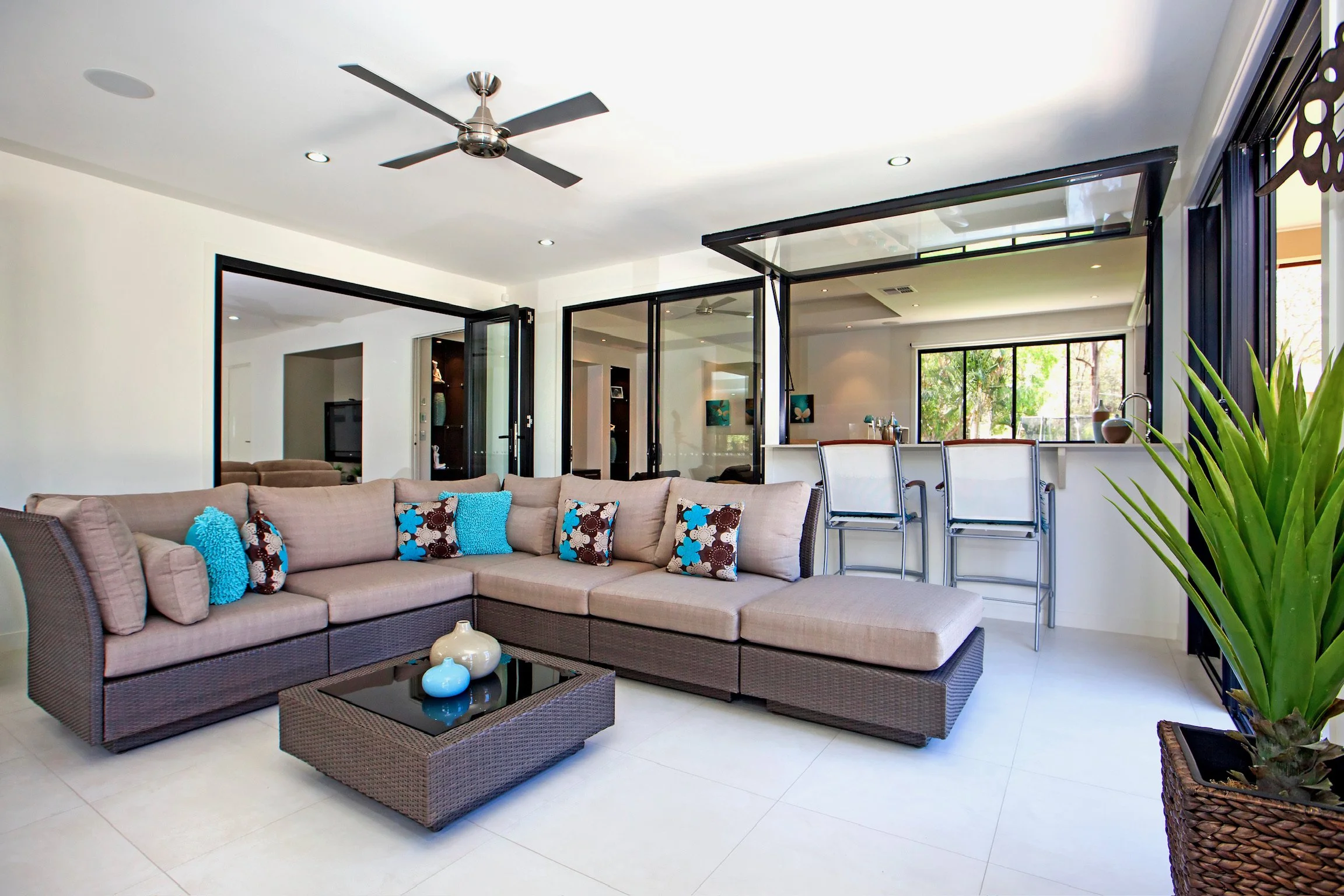 A modern living room with a beige sectional sofa decorated with colorful throw pillows, a glass coffee table with vases, a ceiling fan, and large glass sliding doors leading to an outdoor area with greenery.