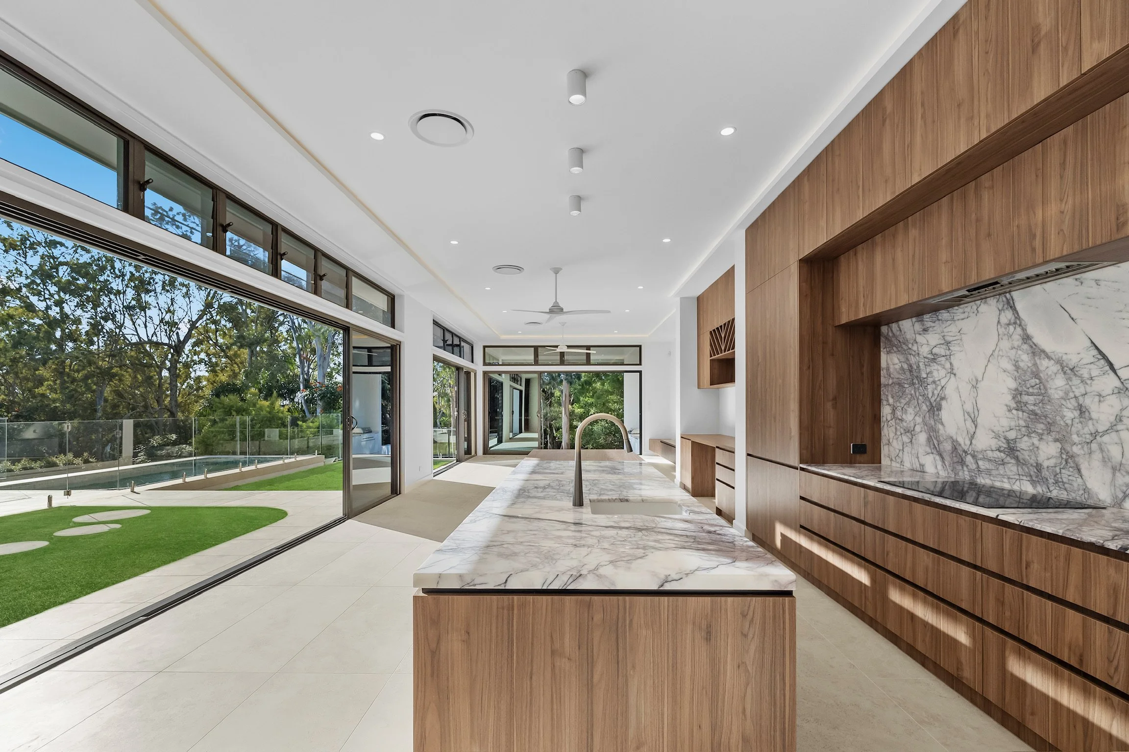 Modern kitchen with marble countertops, wooden cabinets, large glass sliding doors leading to a green backyard, and a ceiling fan.