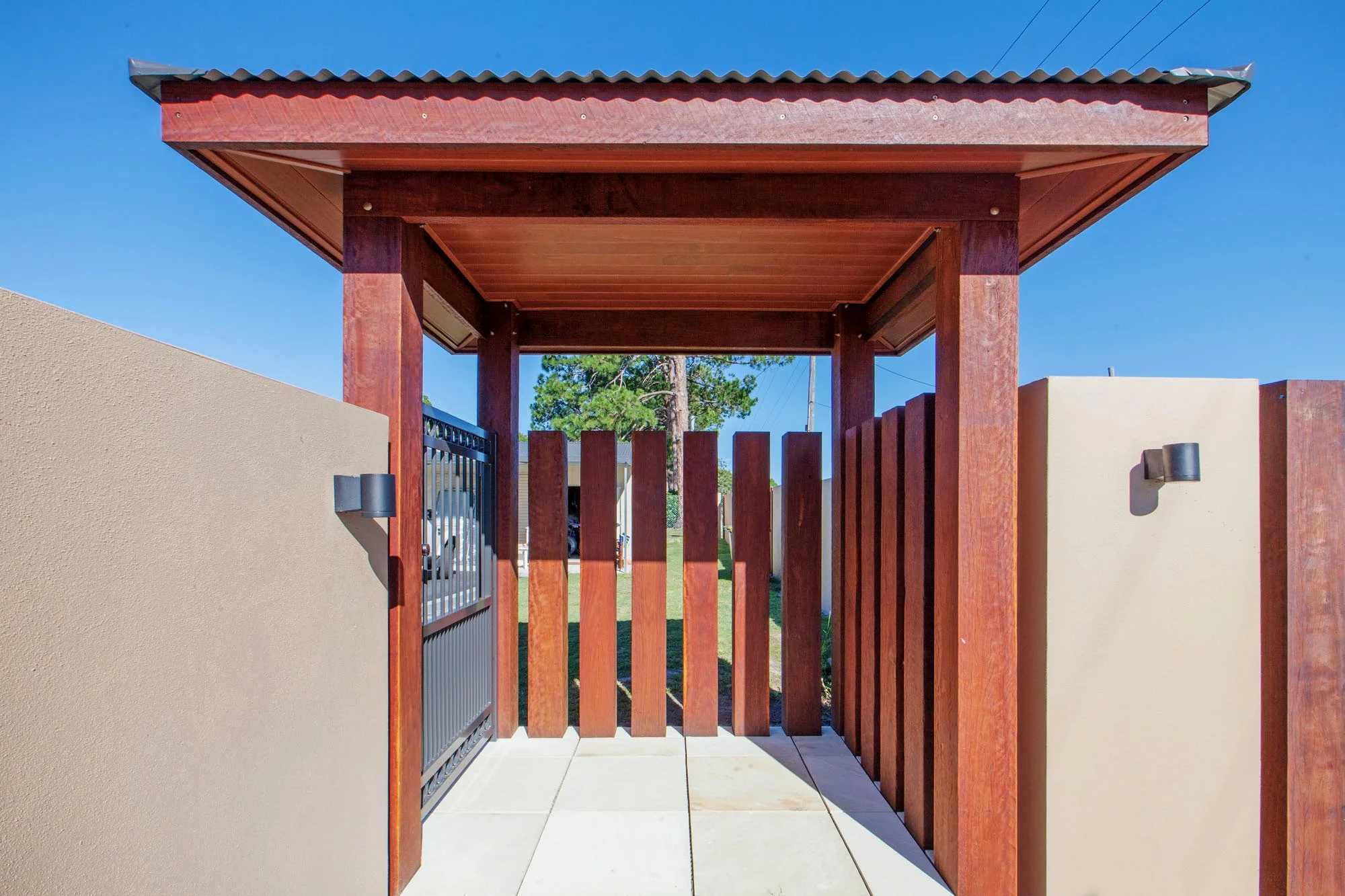 Wooden gate and fence at entrance of yard, flanked by beige wall and metal gate, with a covered top and clear sky in background.