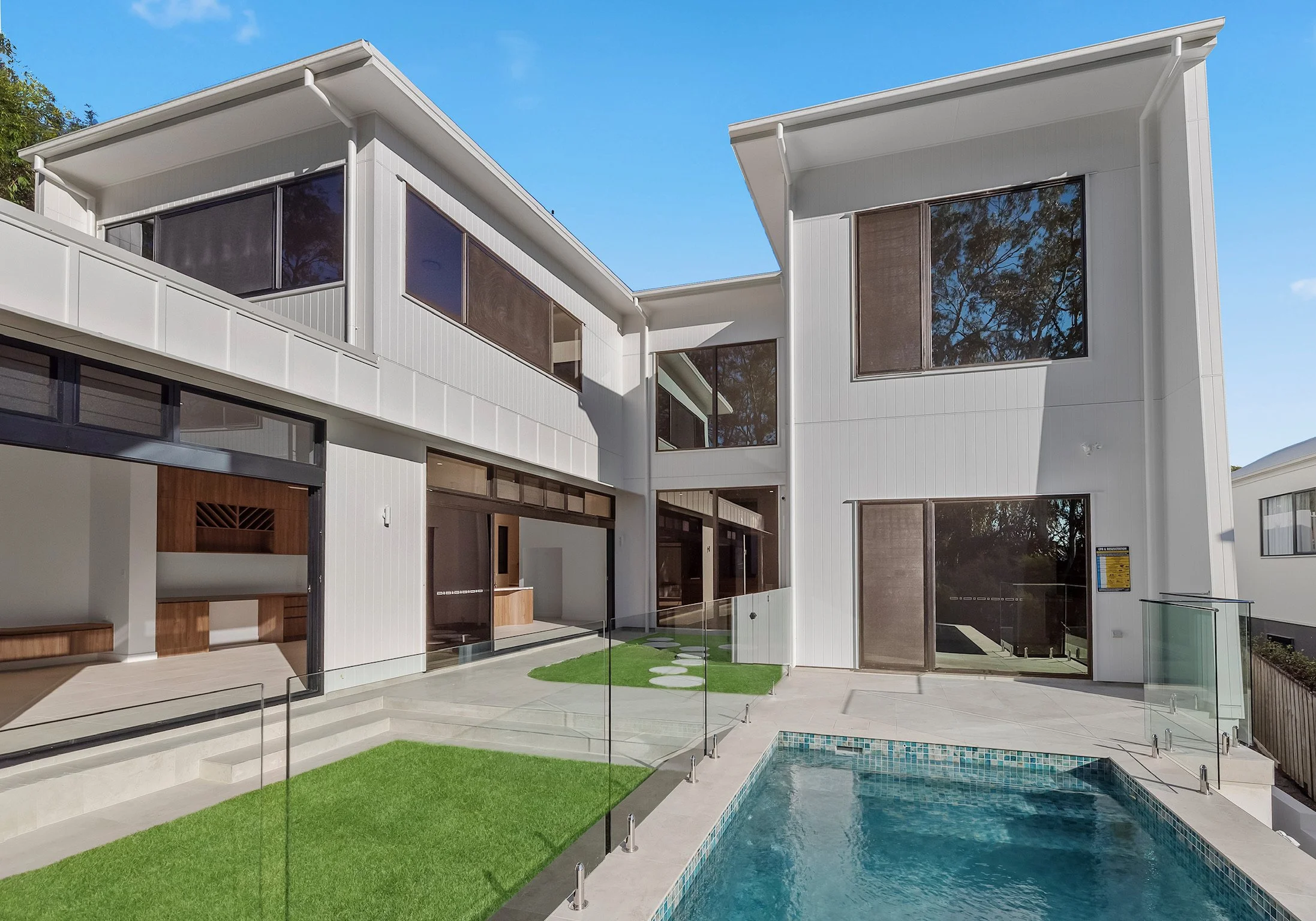 Modern white multi-story house with large windows, small pool, and a yard with artificial grass and stepping stones under a clear blue sky.