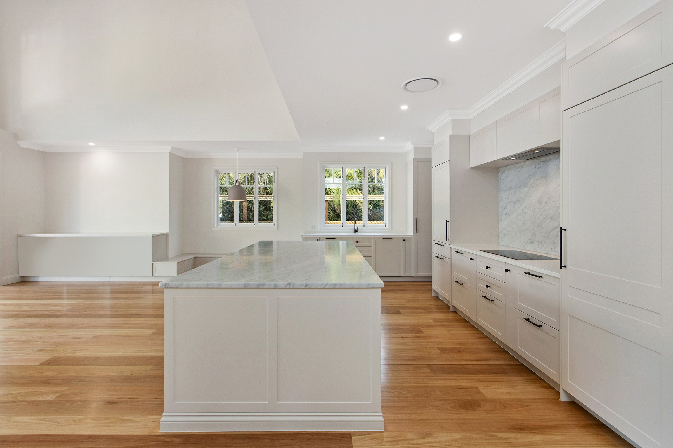 A spacious modern kitchen with white cabinetry, a large marble island, hardwood floors, two windows, and a hanging pendant light.