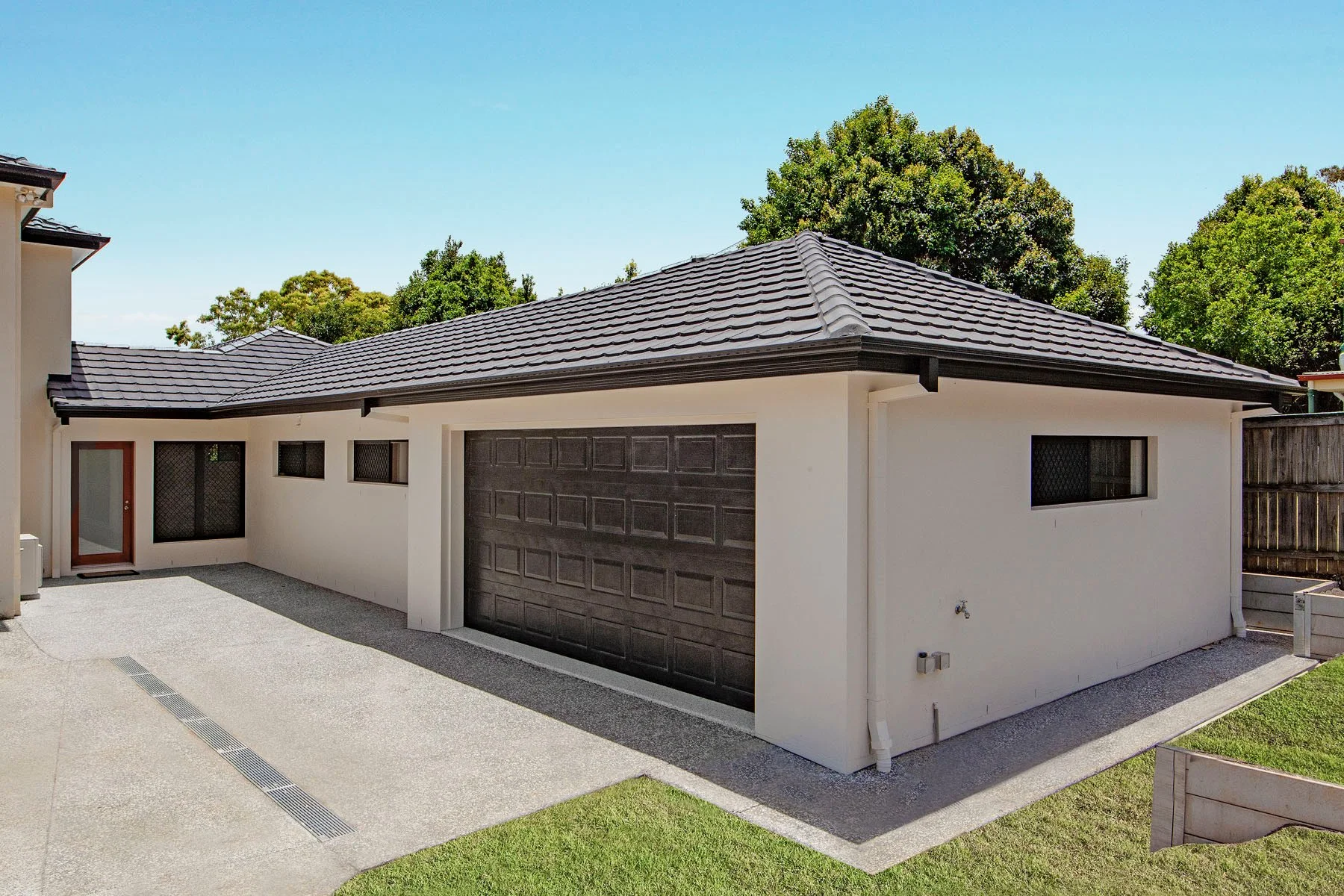 Modern residential house with a dark garage door, white walls, and a tiled roof, surrounded by trees and a concrete driveway.