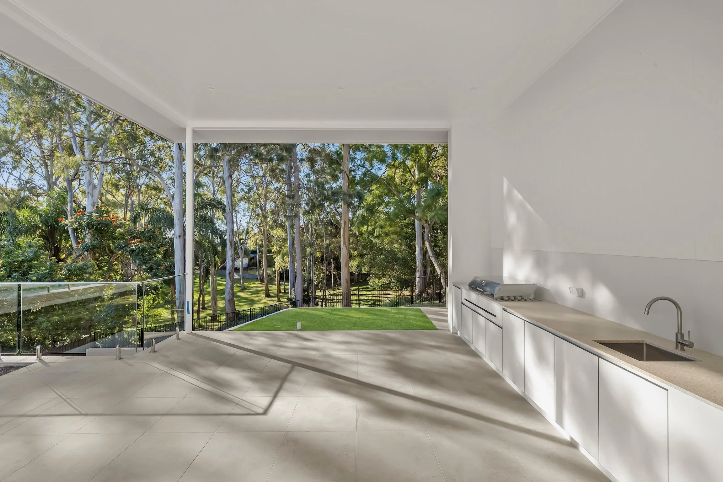 Modern outdoor patio with white cabinetry, a built-in grill, and a view of green trees and blue sky through large open windows