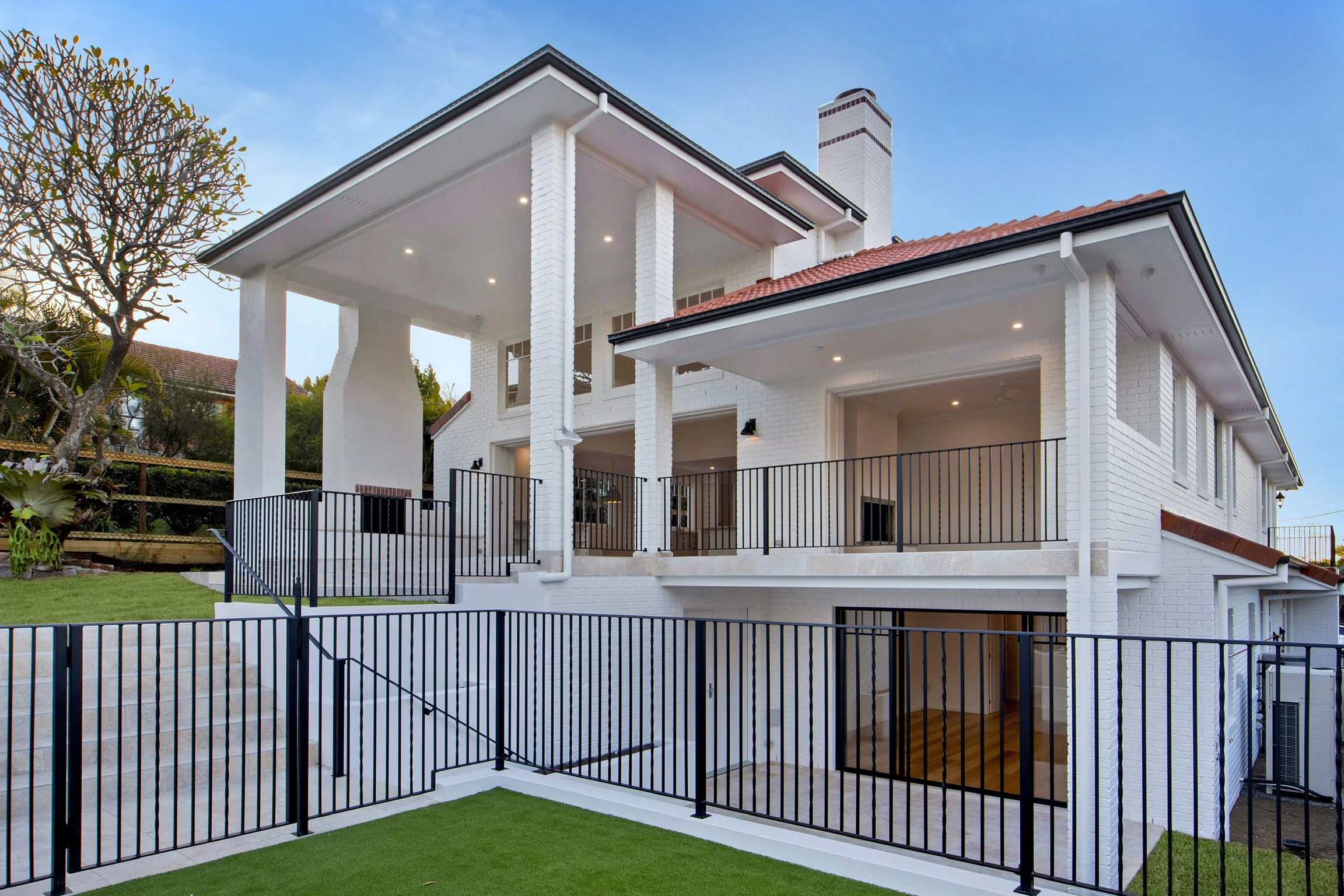 A modern white multi-story house with a brick exterior, multiple balconies, black metal fences, and a well-maintained lawn under a blue sky.