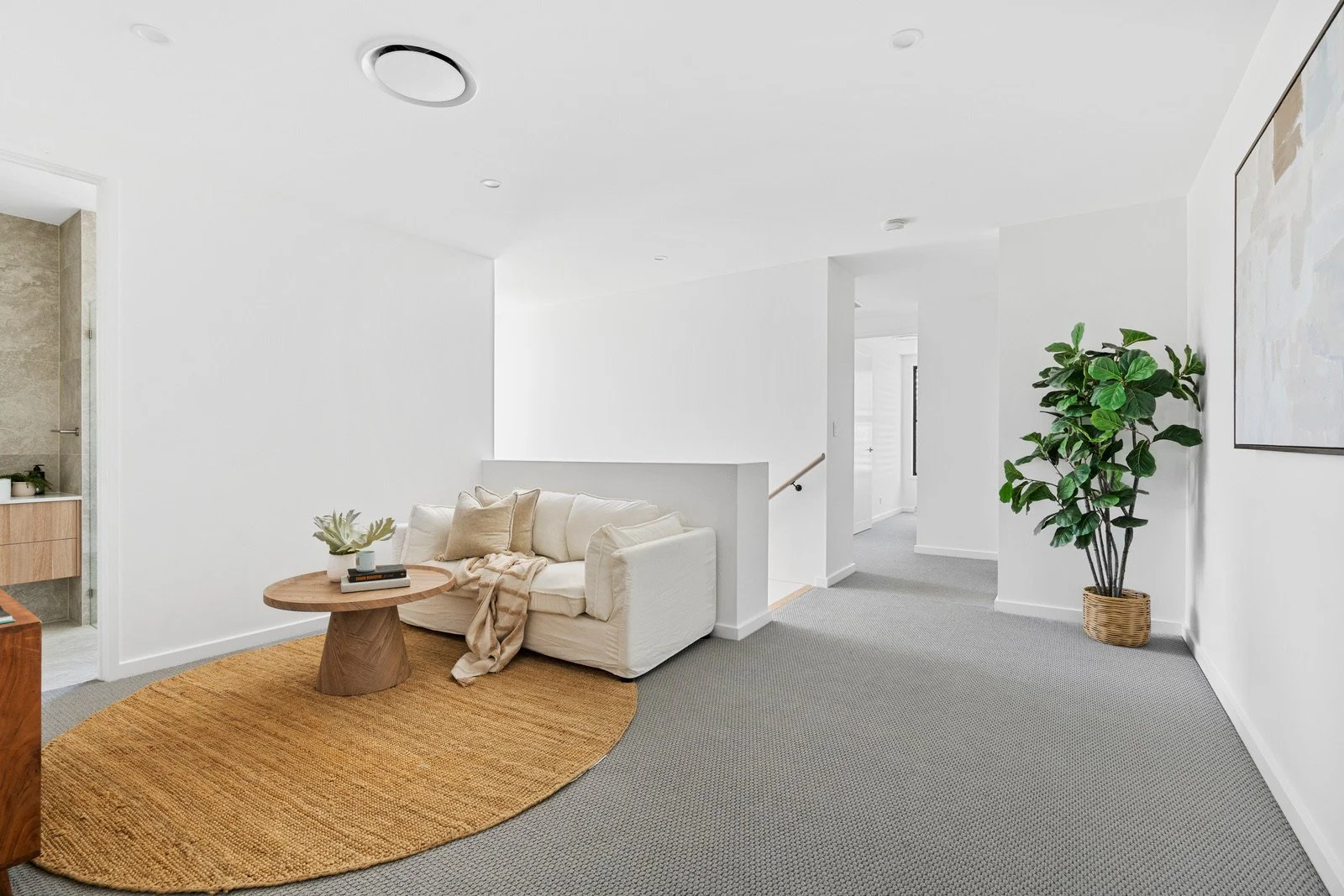 A bright minimalist living room with a white sofa, a wooden coffee table on a tan rug, large potted plant, and artwork on white walls.