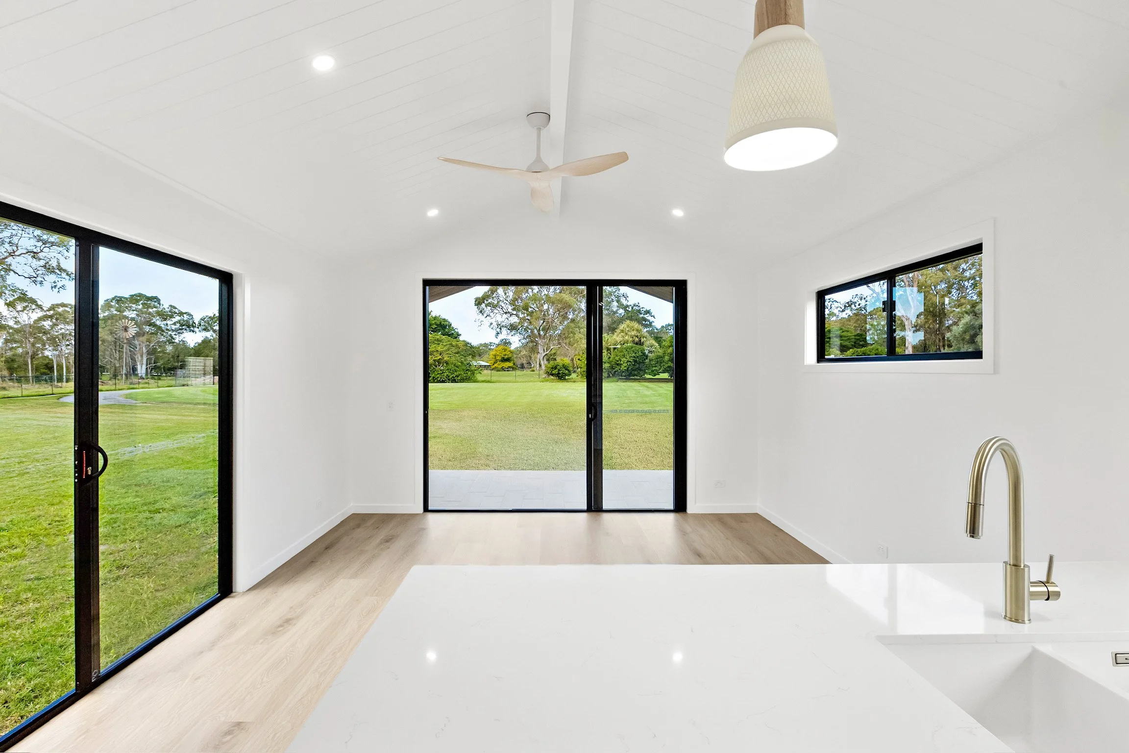 A modern kitchen with white walls, wooden flooring, large sliding glass doors, and windows overlooking a green yard. The ceiling has a white ceiling fan and recessed lighting.