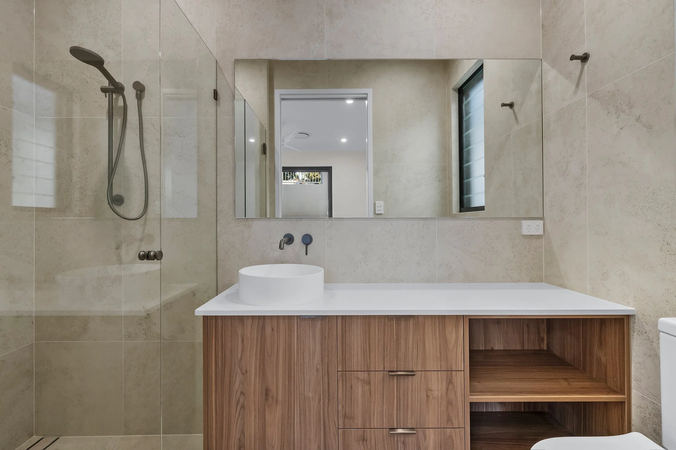 Modern bathroom with beige tiles, a large mirror, a white vessel sink on a wooden vanity, a shower area with a handheld showerhead, and a small window with frosted glass.