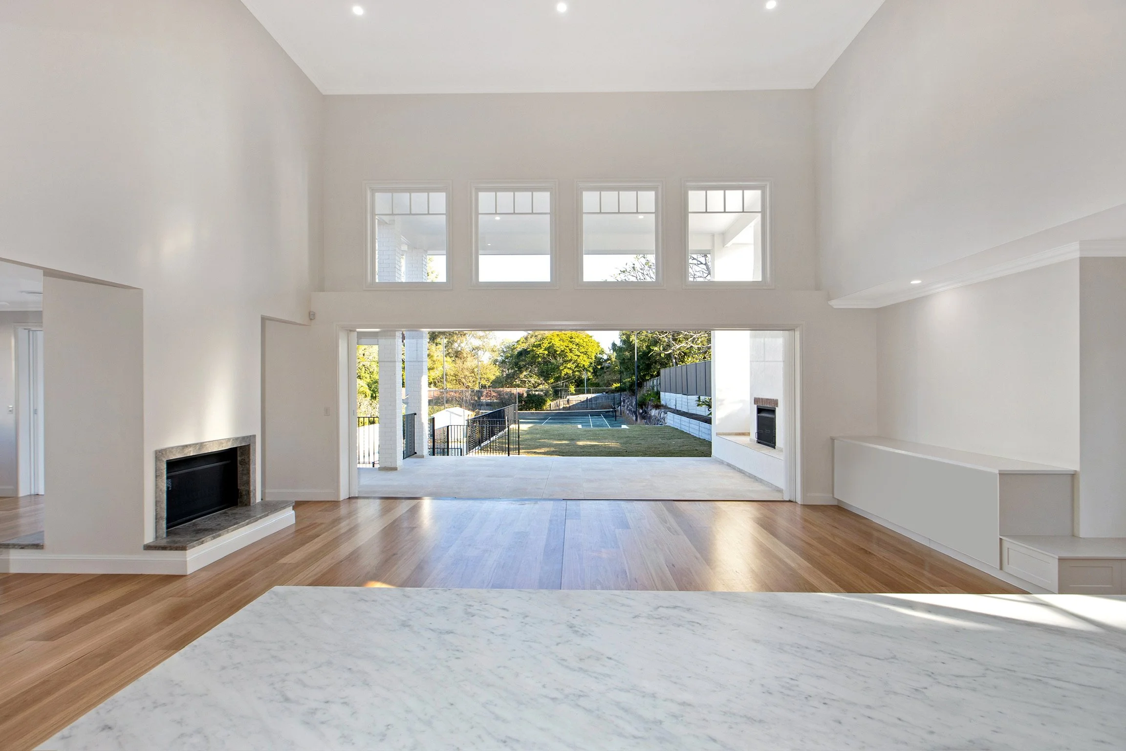 View of a spacious, bright living room with large white walls, high ceilings, a fireplace, hardwood floors, and large windows looking out to a backyard with a tennis court surrounded by trees.
