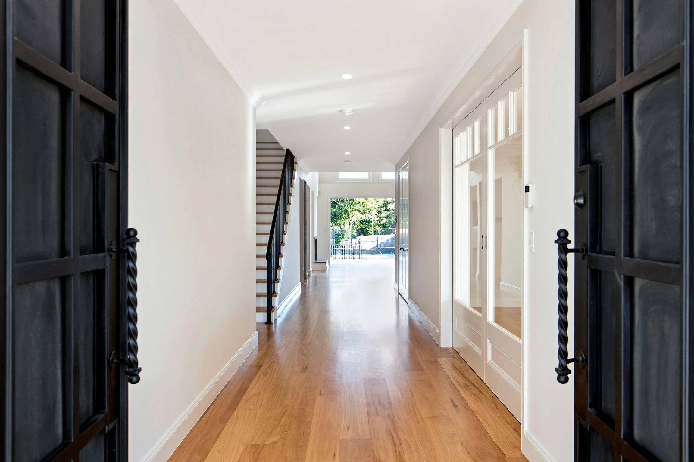 View of a bright hallway seen through open black iron gates, with hardwood flooring, white walls, staircase, and windows in the background.