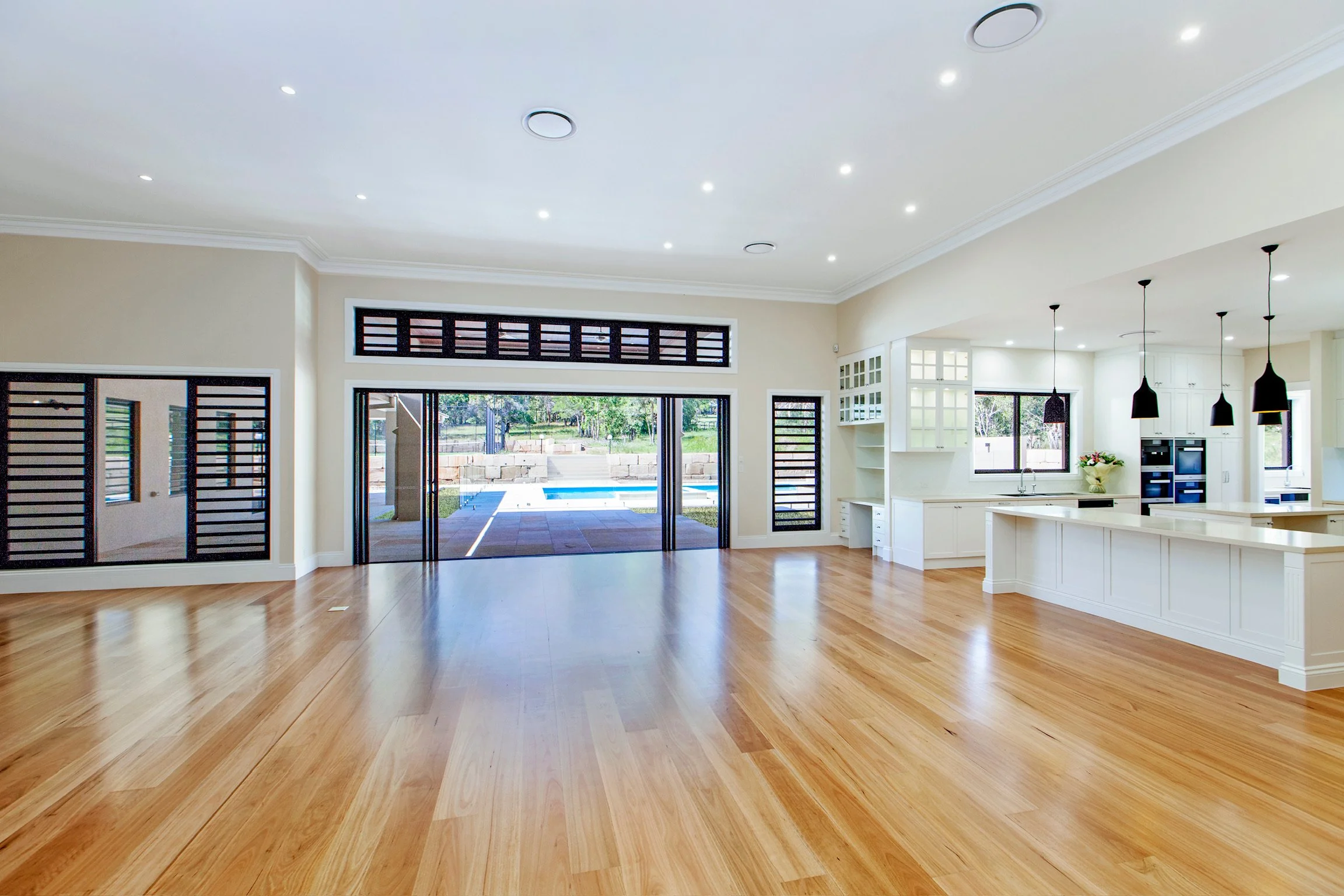Open-concept living space with wooden flooring, large windows, and a view of a backyard with a swimming pool, connected to a white kitchen with black pendant lights.