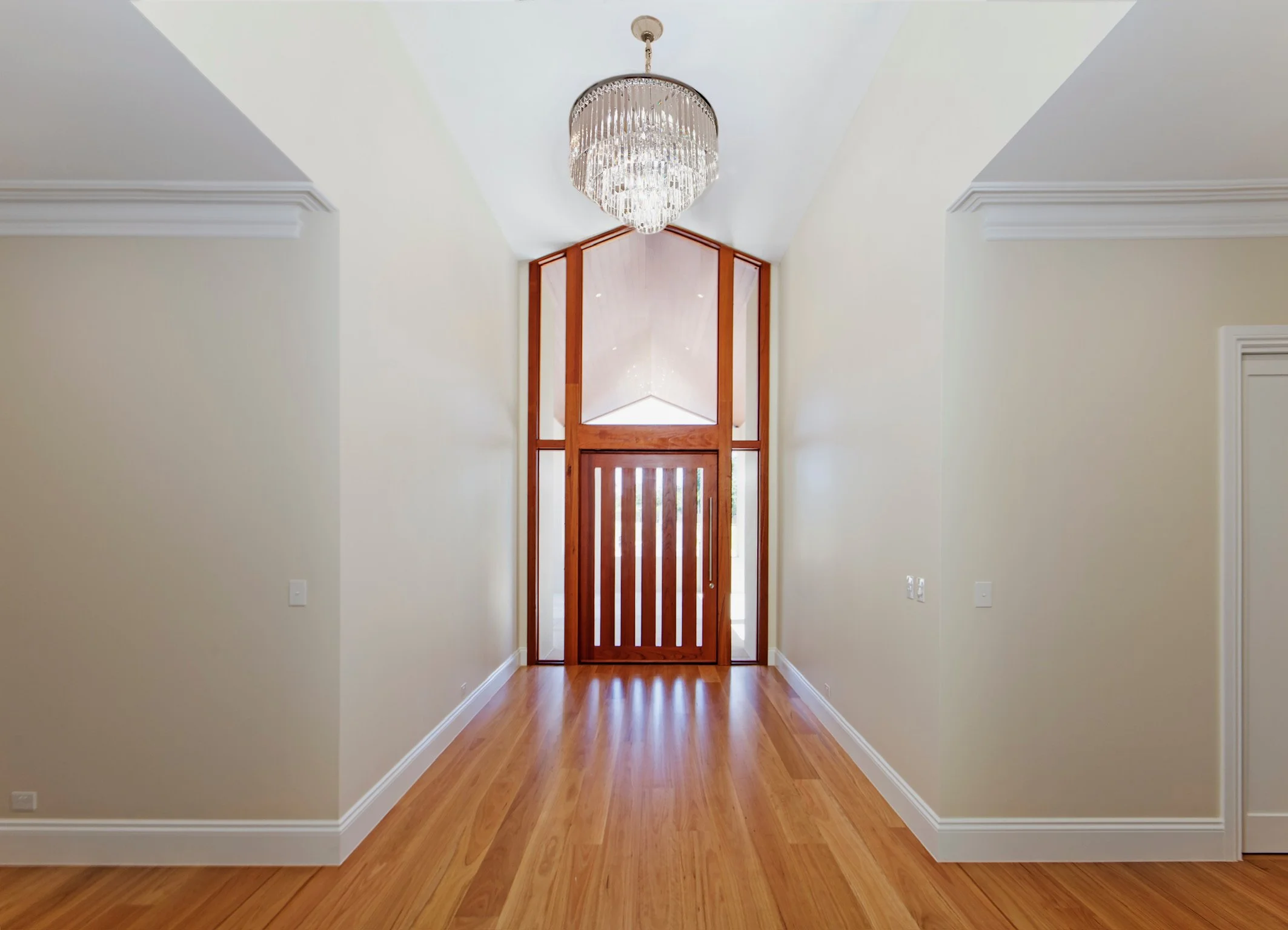 A hallway with light-colored walls, wooden flooring, crown molding, and a wooden front door with glass panels. A chandelier hangs from the ceiling.