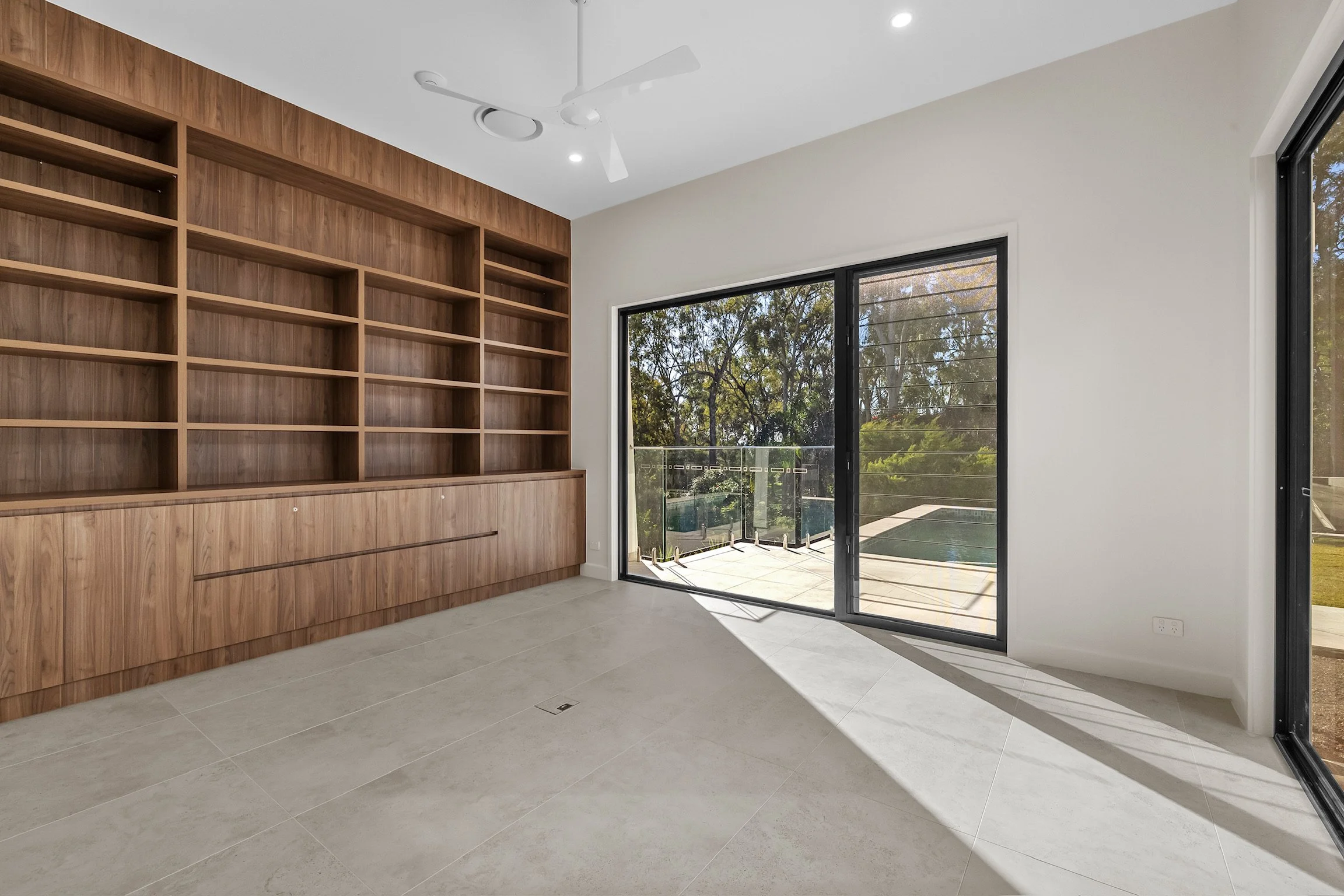 Empty room with wooden built-in shelves, large sliding glass doors leading to an outdoor patio, and trees visible outside.