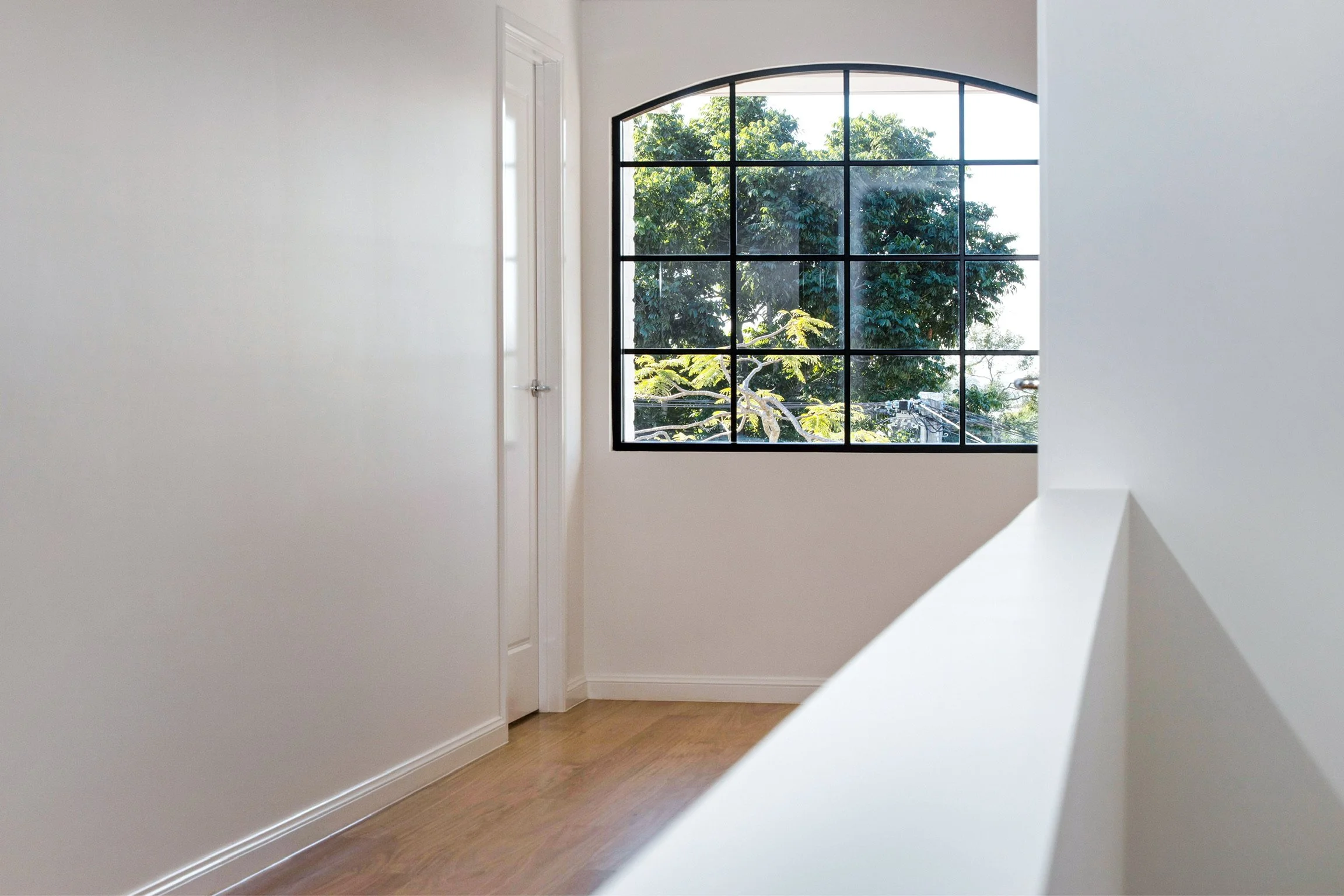 Interior view of a room with a large arched window showing green trees outside, white walls, a white door, and wooden flooring.