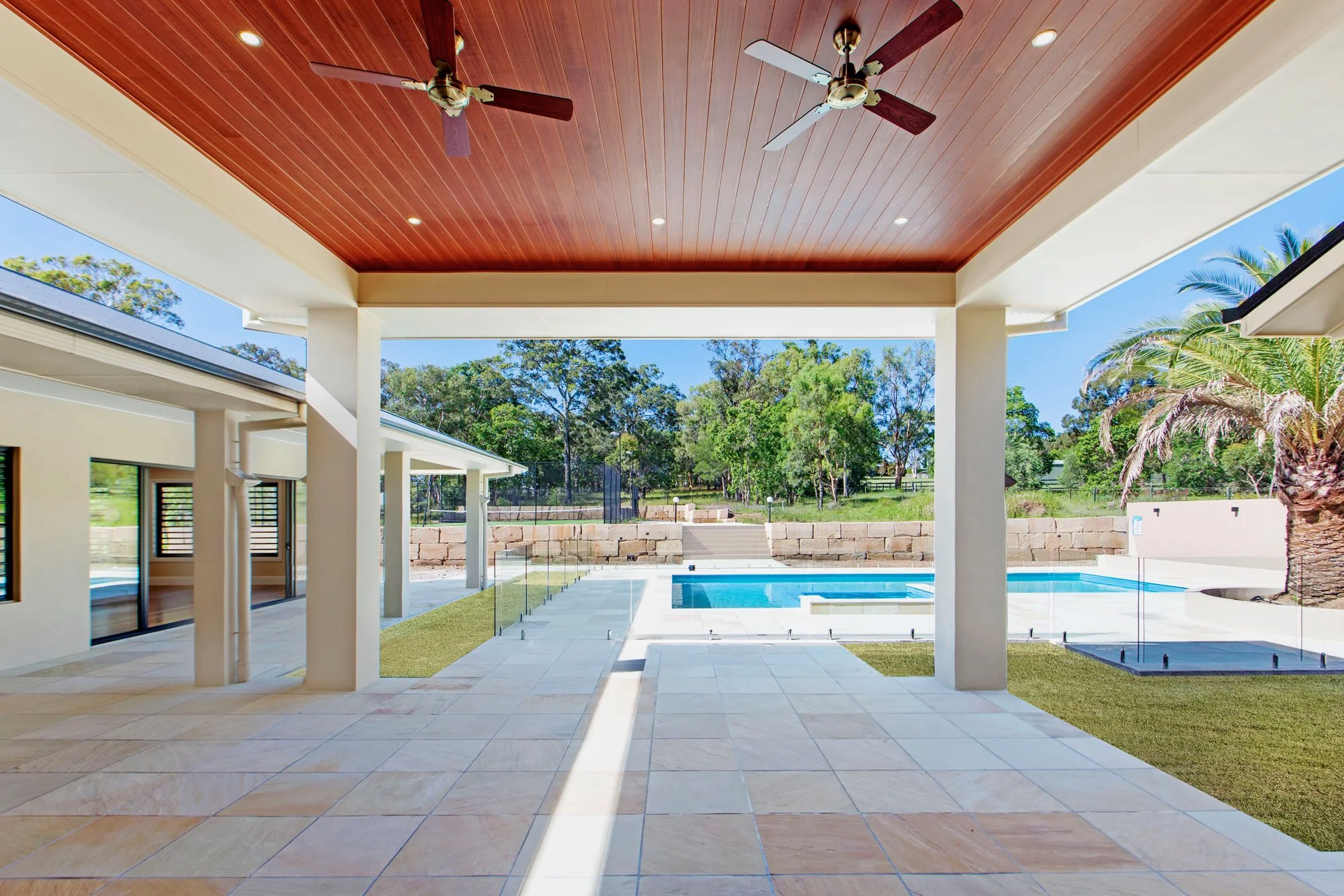 Covered patio area with ceiling fans, recessed lighting, and tile flooring. Beyond, a swimming pool and a landscaped yard with trees and a brick wall in the background.