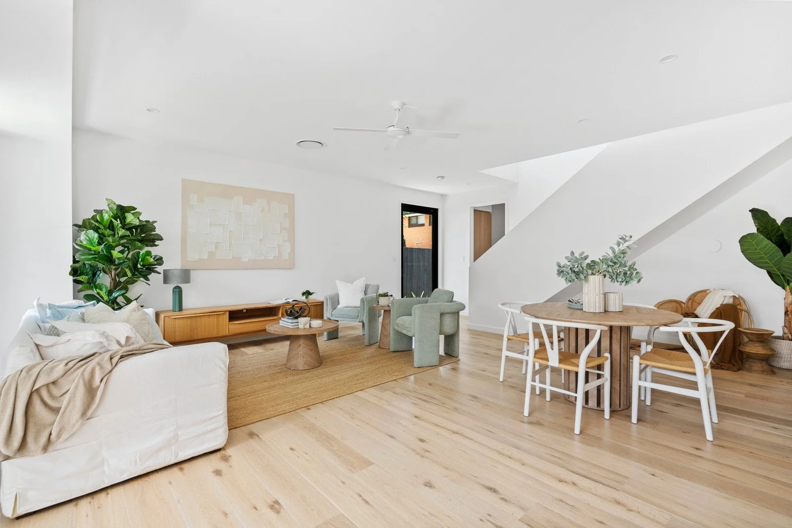 Living and dining room with white walls, light wood flooring, and modern furniture. A beige sofa with pillows, a green armchair and side table, a wooden speaker, and a large green plant on the left. A round wooden dining table with white chairs, a la
