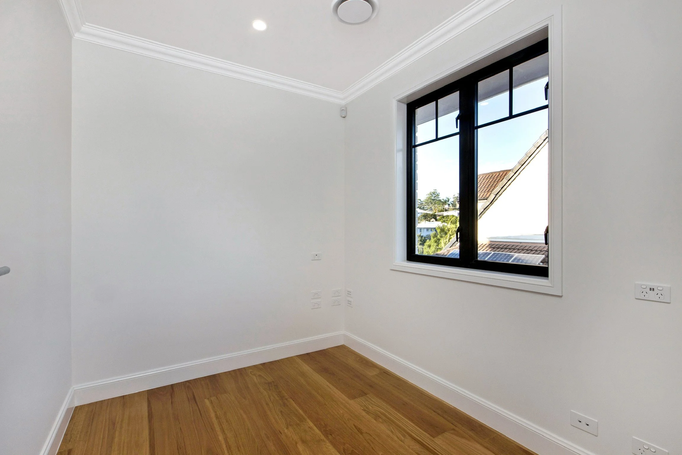 Empty room with a large window, white walls, and wood floors. Outside the window, trees and rooftops are visible.