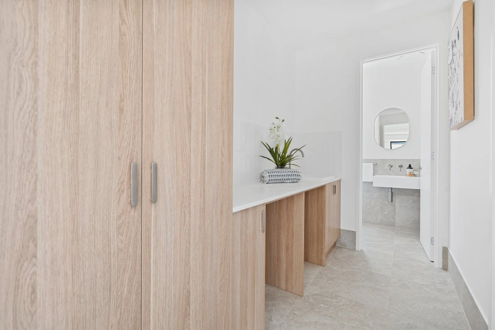 A minimalist bathroom with a white vanity, oval mirror, and a potted plant on a wooden cabinet. Neutral tones and natural wood accents.