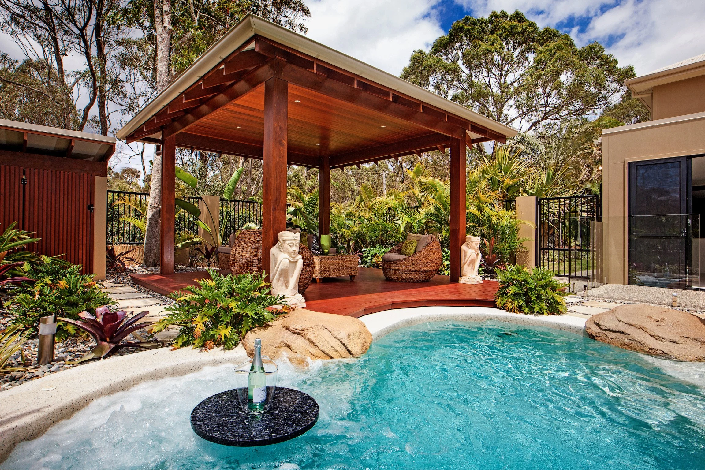 A backyard with a swimming pool, outdoor seating area with wicker chairs, statues, lush greenery, and trees under a blue sky.
