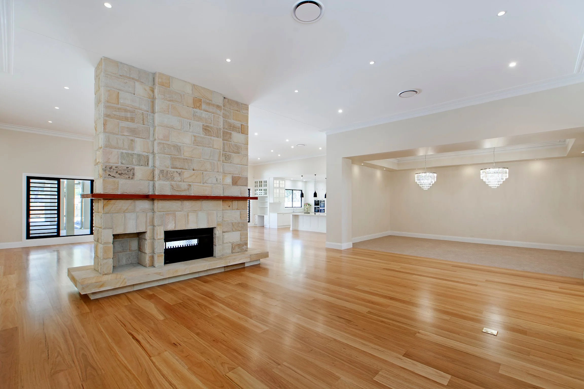 Interior of a spacious, modern living room with hardwood floors, a stone fireplace, and high ceilings with recessed lighting. There are chandeliers hanging in the background and multiple windows providing natural light.