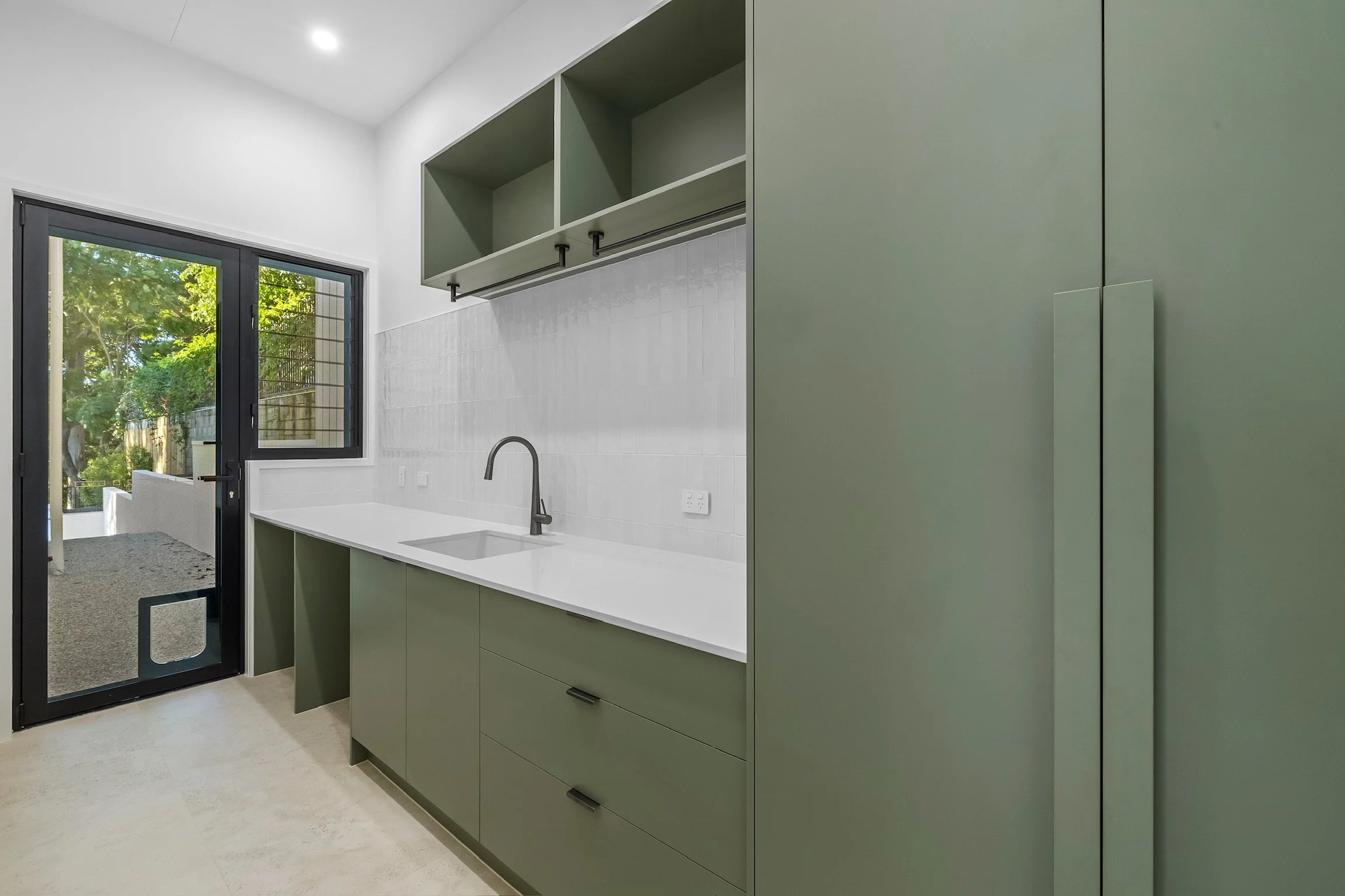 Modern kitchen with green cabinets, a white countertop, a black faucet, white tiled backsplash, a large window showing trees outside, and a sliding glass door leading to an outdoor area.