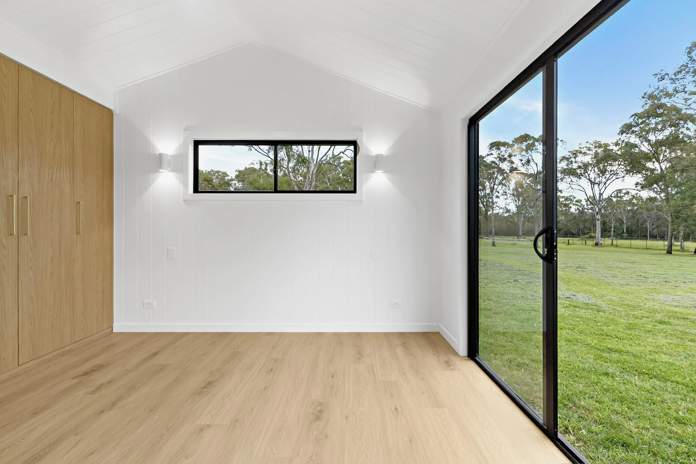 A modern interior room with white walls, tall wooden closet doors, a small horizontal window, and large sliding glass doors revealing a green outdoor landscape with trees and a fence.