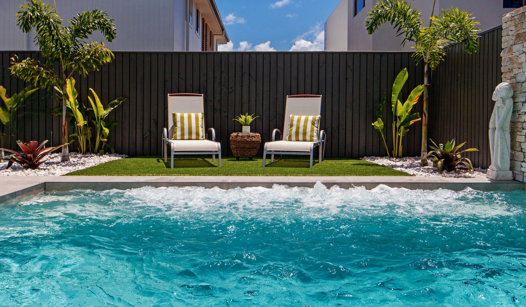 A backyard swimming pool with clear blue water, two white lounge chairs with yellow and white striped pillows, a small potted plant on a wicker table between the chairs, tropical plants, and trees against a black wooden fence, a statue of a woman on 