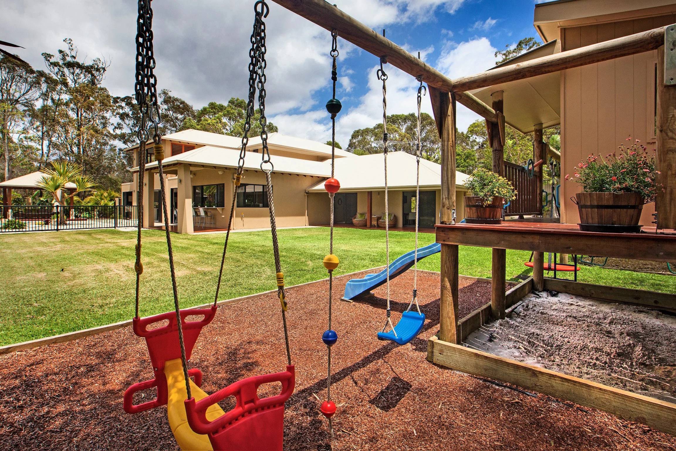 Kids' playground with swings, a slide, and a sandbox, surrounded by a grassy yard and houses with trees and a partly cloudy sky in the background.