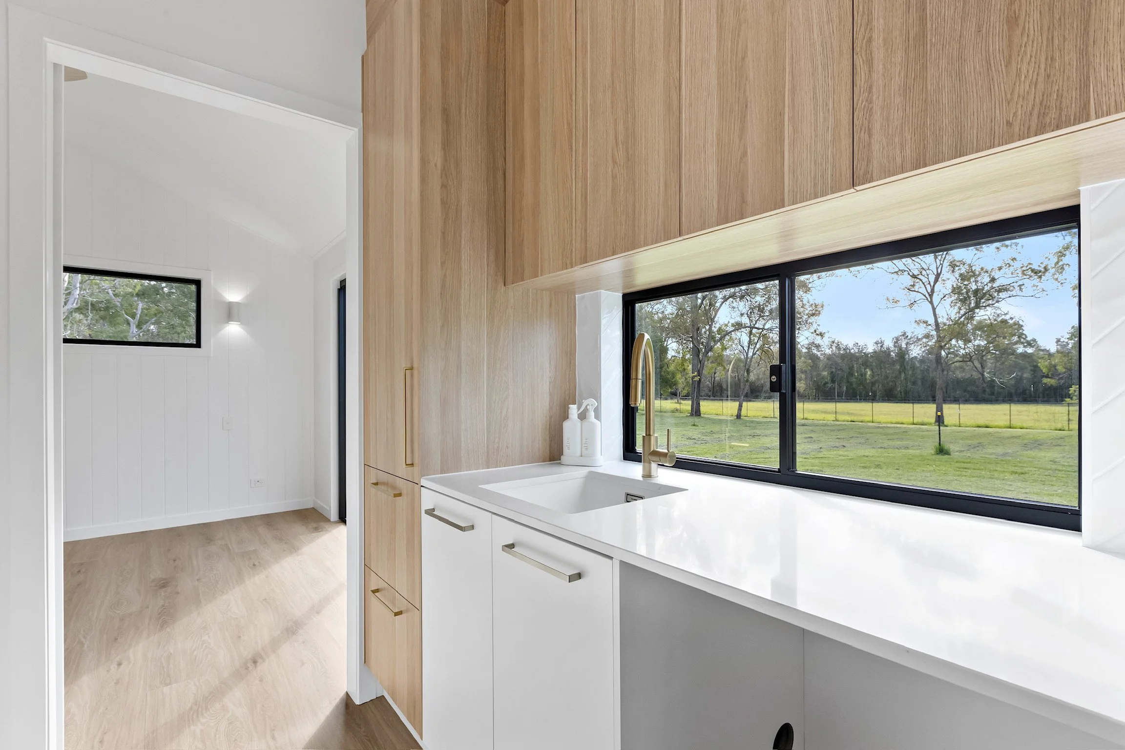 Modern kitchen with white cabinets, a large window showing outdoor greenery, and a white countertop with a sink and golden faucet.