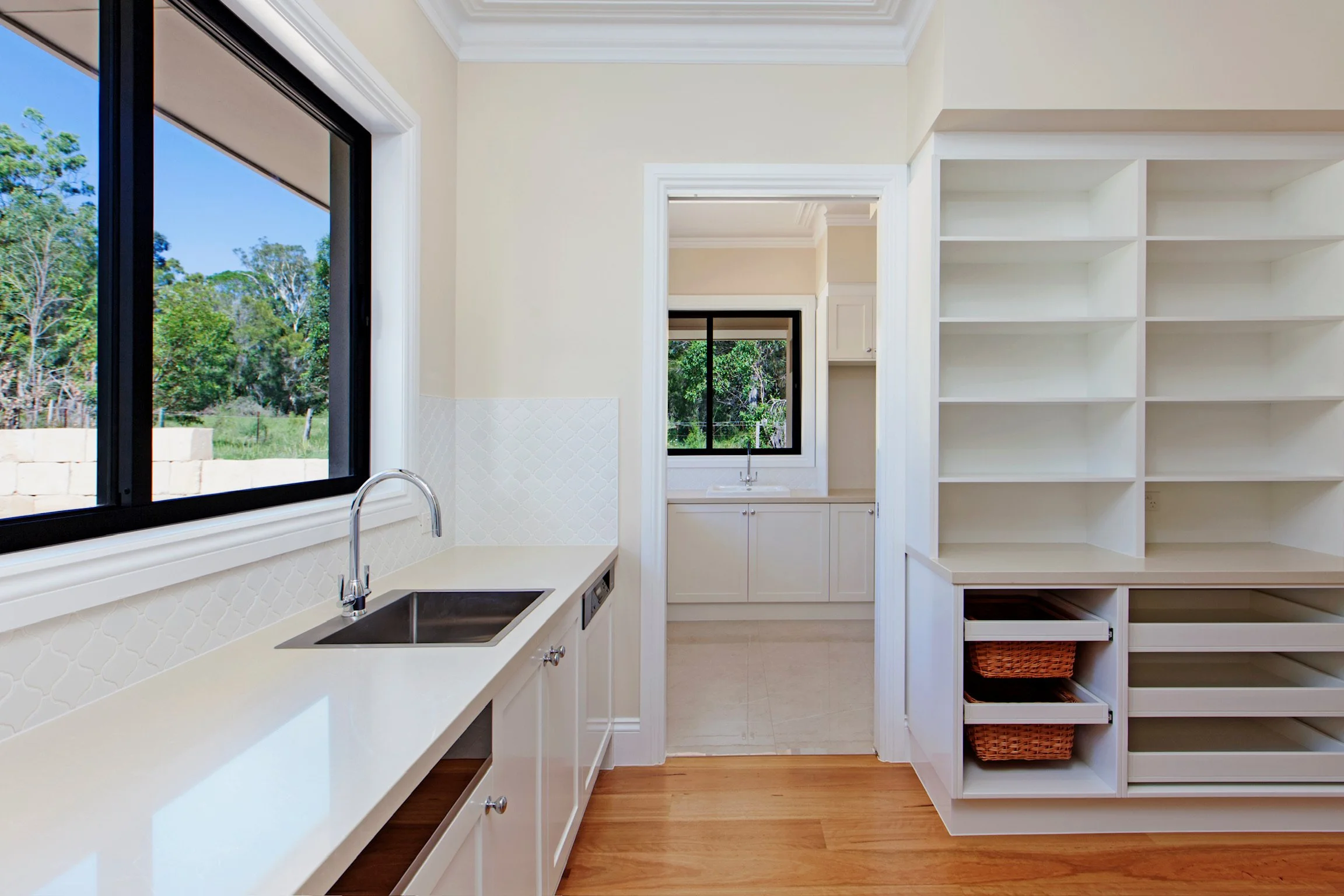 Bright kitchen with large window showing green trees outside, white cabinets, a white countertop with sink and small stove, open shelving with baskets, and polished wood flooring.