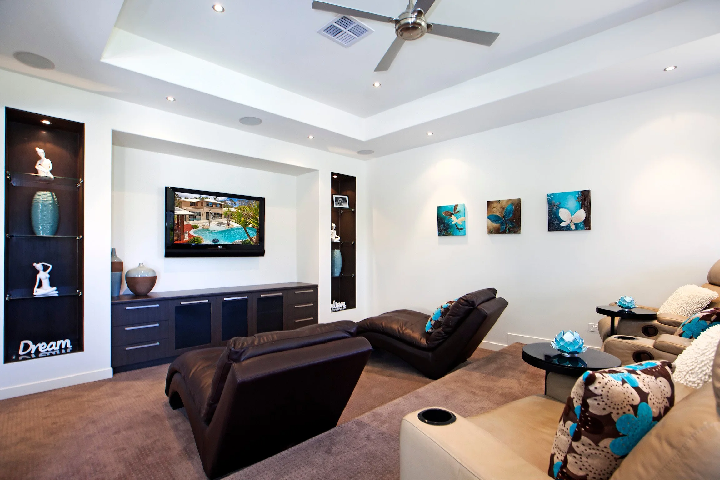 Living room with brown leather sectional sofa, wall-mounted TV showing a pool and house, decorated with blue and white floral accents, abstract butterfly artwork on the white wall, ceiling fan, built-in shelves with decorative items, and a beige otto
