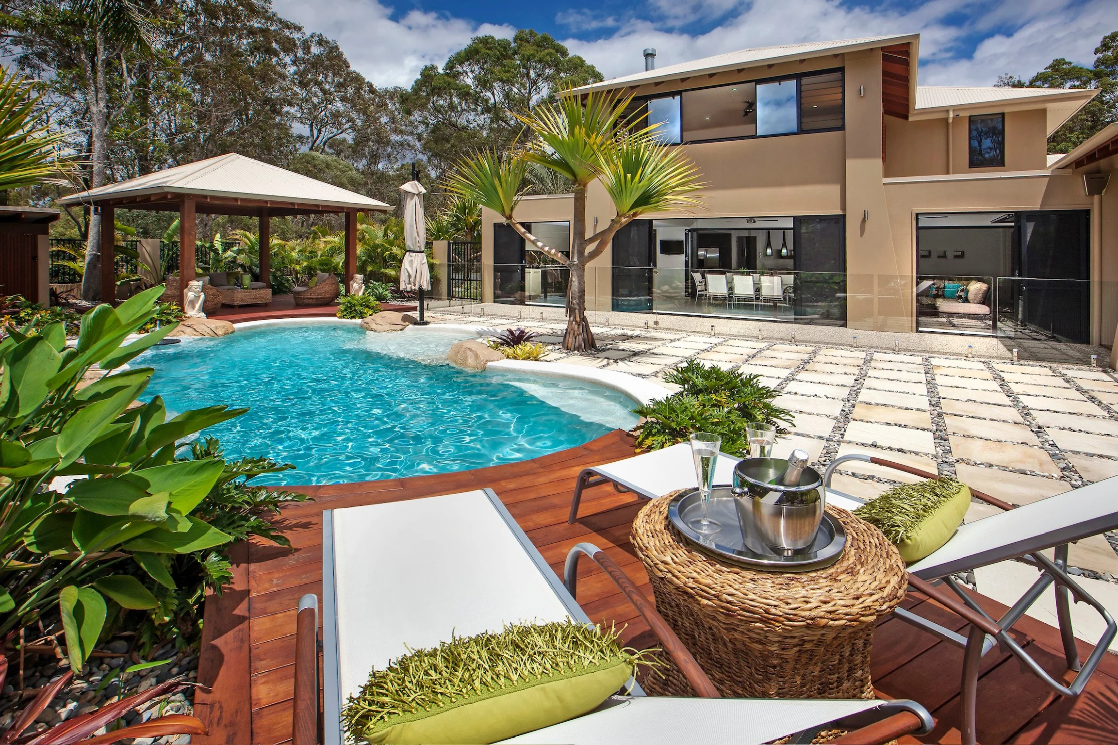 A backyard view of a modern house with large windows, a swimming pool with rocks, lush green trees, and outdoor furniture including lounge chairs, pillows, and a table with drinks and a wine bottle.