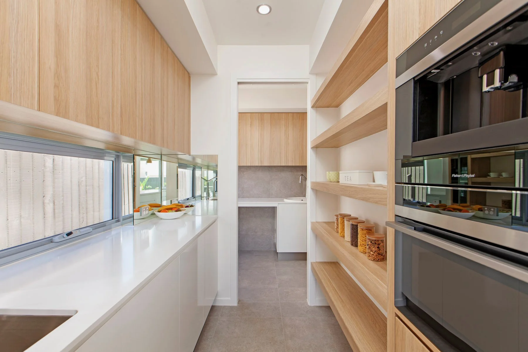 Modern kitchen with white countertops, wooden cabinets, open shelves, and built-in appliances. There are fruit bowls on the counter and jars of spices on the open shelves.
