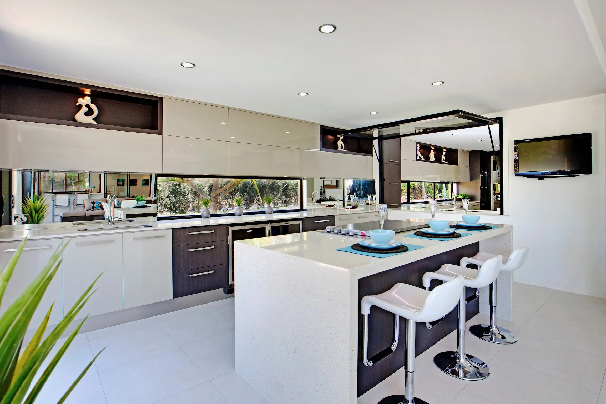Modern kitchen with white countertops, dark cabinetry, large windows, and a kitchen island set with blue bowls, black placemats, and white bar stools.