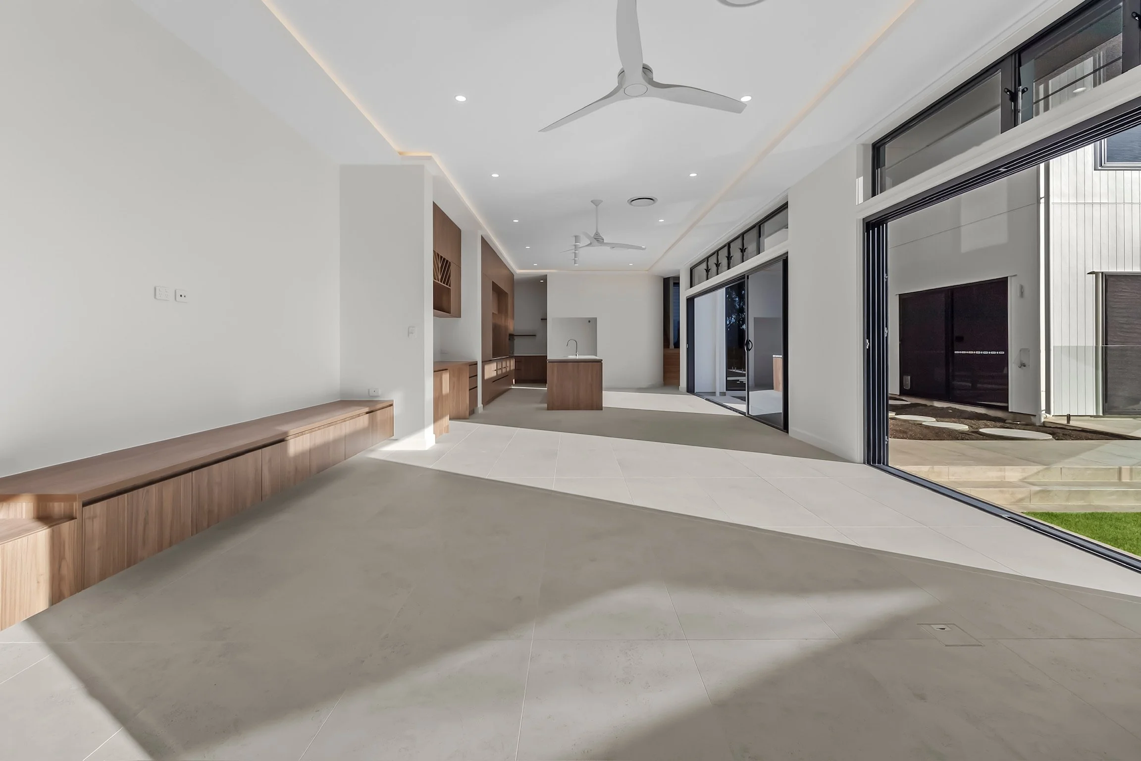 Modern interior living space with white walls, beige tiled floor, wooden built-in cabinetry, large sliding glass doors leading outside, and ceilings with ceiling fans and recessed lighting.
